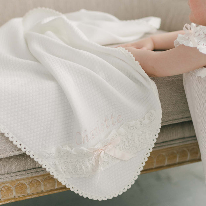 A baby is lying on a beige couch, partially covered by the Charlotte Personalized Blanket, an ivory quilted cotton blanket featuring lace edging. This keepsake is embroidered with the name "Charlotte" in soft pink thread and adorned with a delicate pink silk ribbon bow.