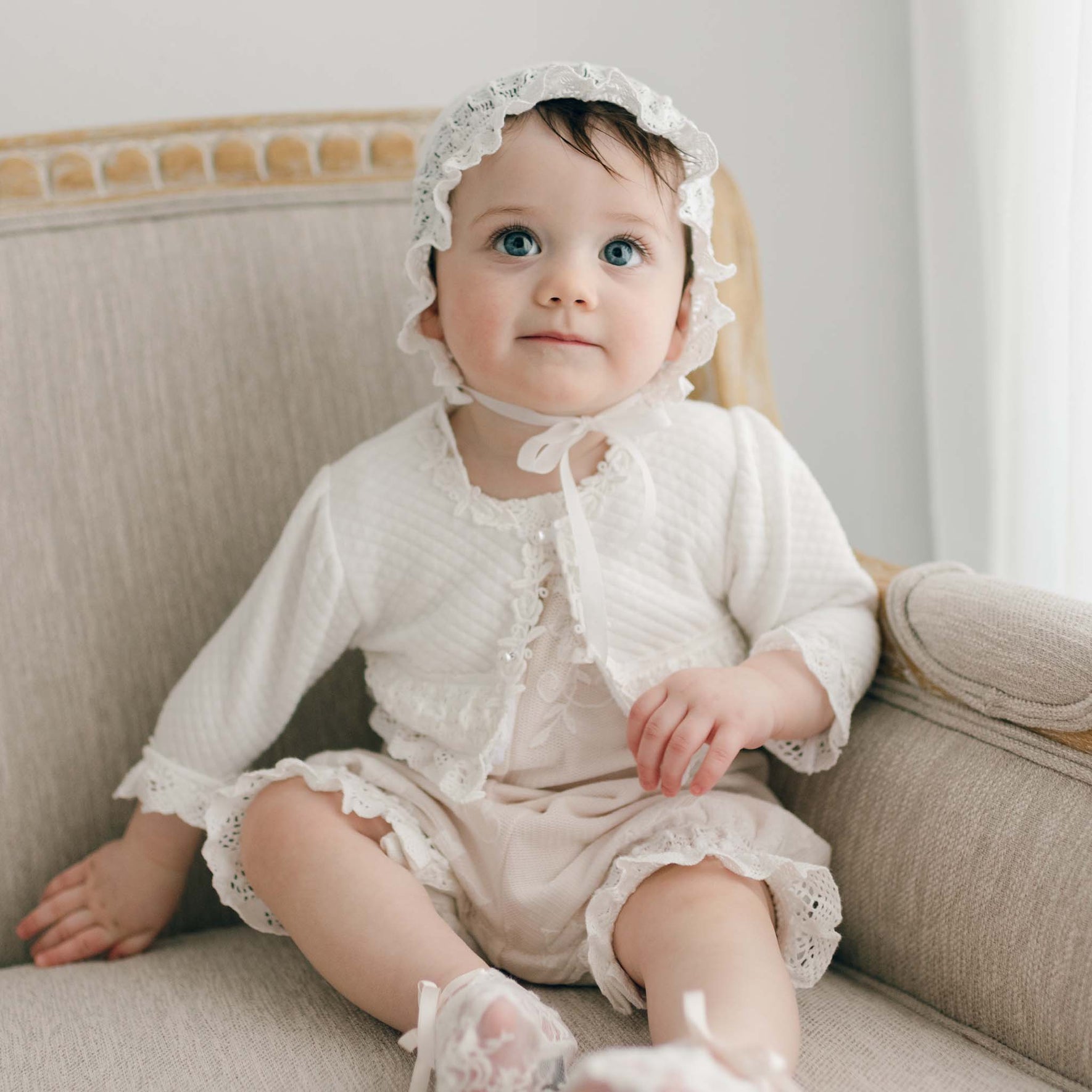 A baby sits on a beige armchair wearing the Charlotte Quilted Cotton Bonnet, featuring a white lace-trimmed design with an outfit that matches its handcrafted ivory quilted cotton material. The baby gazes upward with a neutral expression as the armchair's light upholstery and wooden details contribute to the softly lit, gentle atmosphere.
