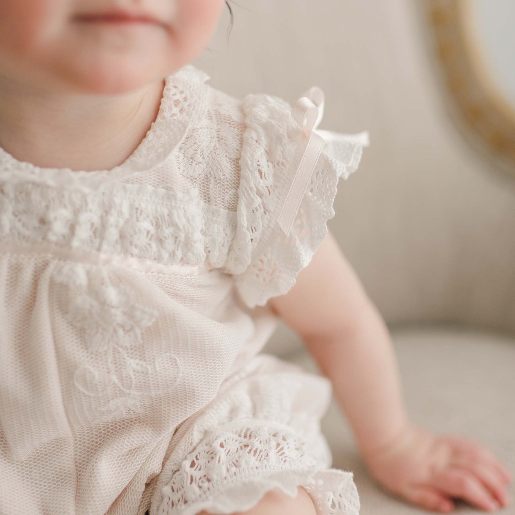 Close-up of a toddler in a delicate Charlotte Romper with lace-trimmed details, sitting on a light-colored cushioned chair. The focus is on the intricate lace accents against the softly blurred background, highlighting its elegance and charm.