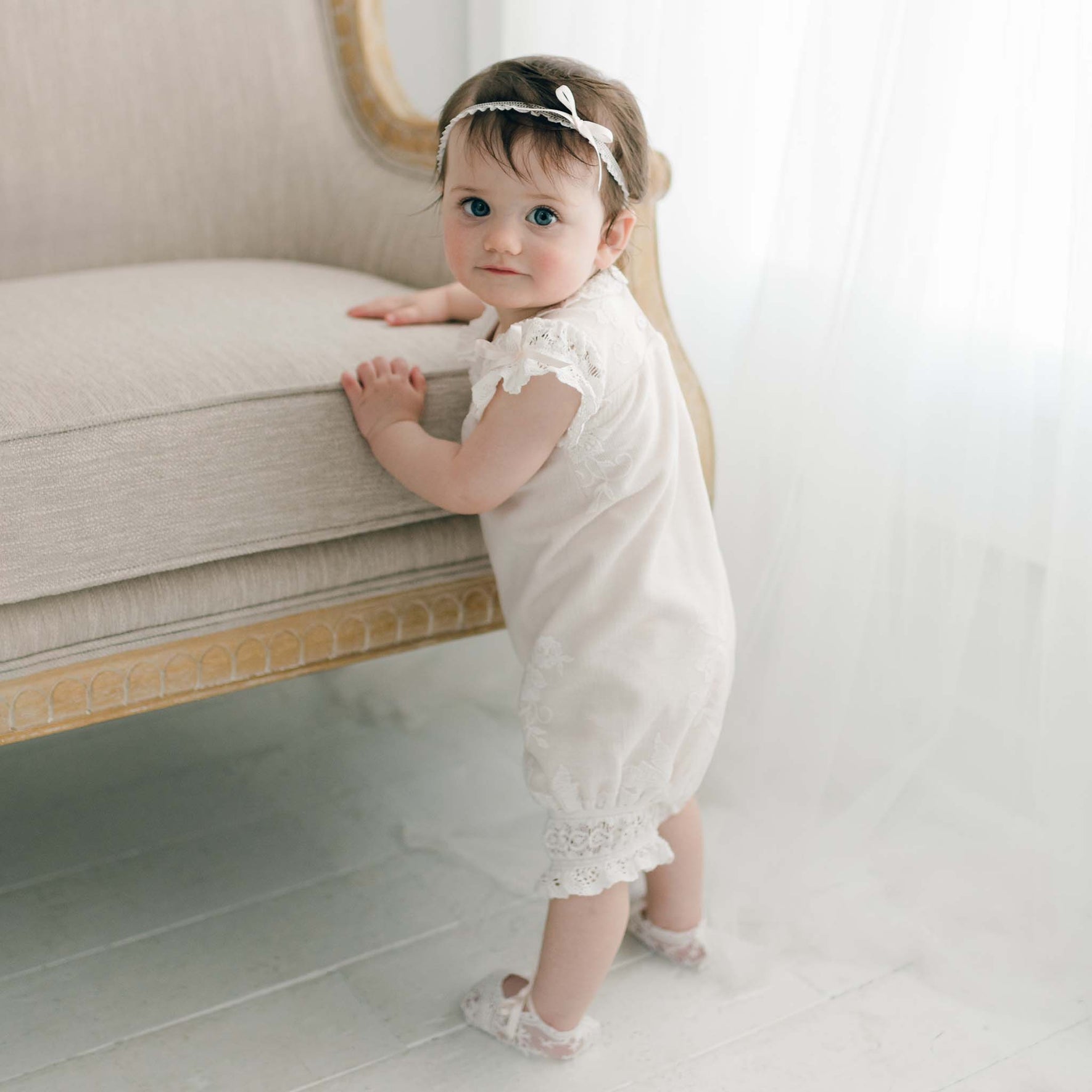 A baby stands holding onto a beige armchair, dressed in the Charlotte Romper and matching headband. The room is softly lit with a sheer curtain in the background. The embroidered cotton lace details of the romper catch the light as the baby looks over their shoulder toward the camera.