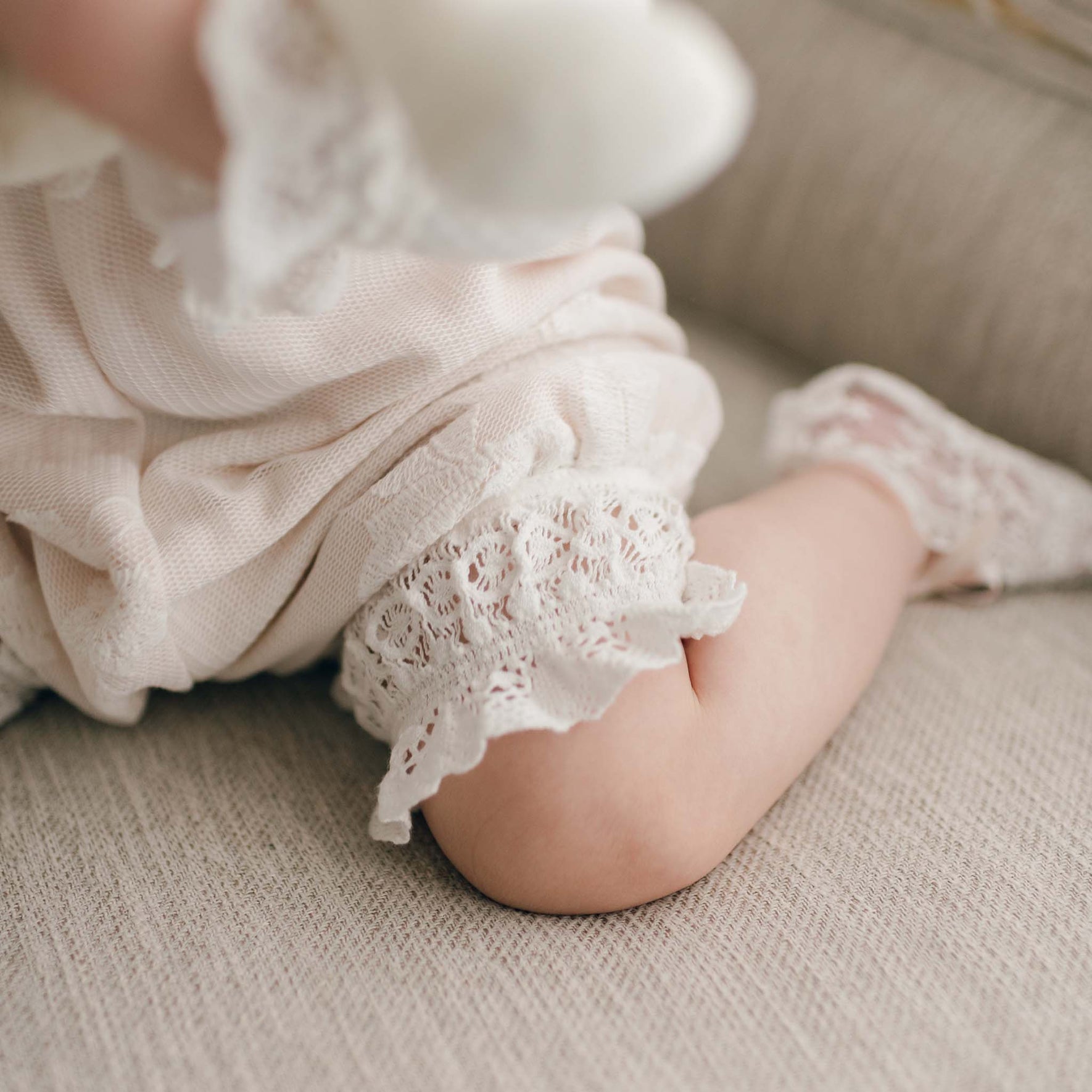 A close-up of a baby's leg in the Charlotte Romper, a cream-colored Christening outfit adorned with embroidered cotton lace trim and lined with pima cotton. Tiny socks rest on the beige fabric surface, while the baby's arm peeks through, showcasing delicate lace details on the sleeve.