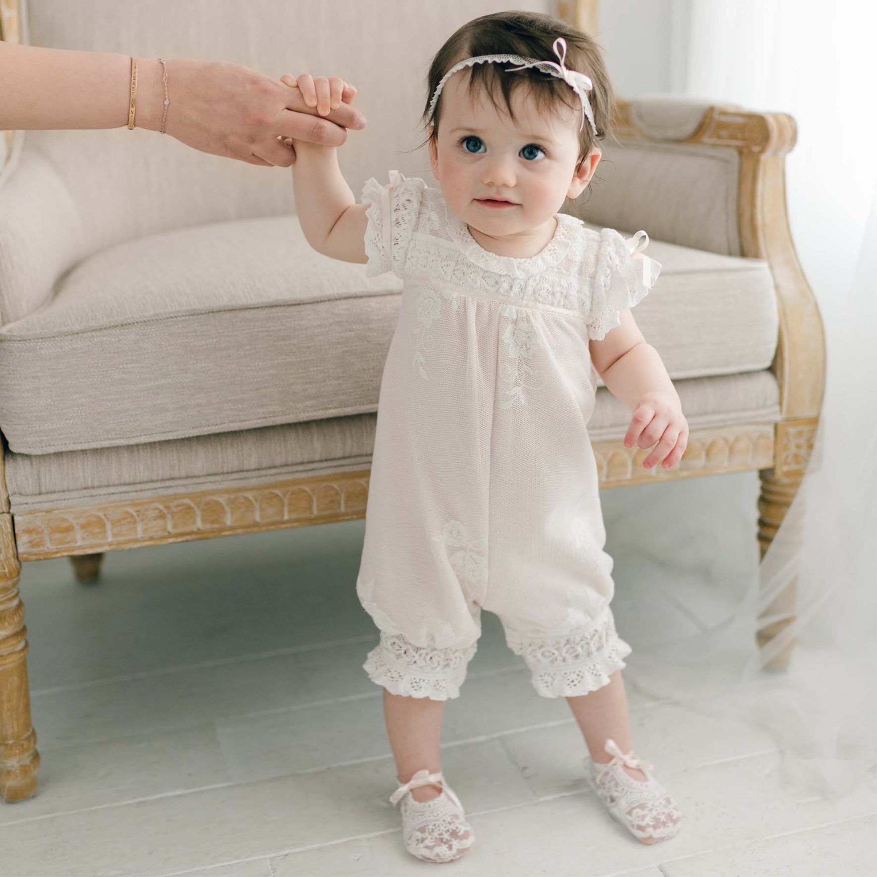 A baby wearing the Charlotte Romper, made of white embroidered cotton lace, stands on a white floor while holding an adult's hand. The baby is adorned with a headband featuring a bow and stands in front of a light-colored, ornate chair. The softly lit setting adds to the gentle atmosphere.