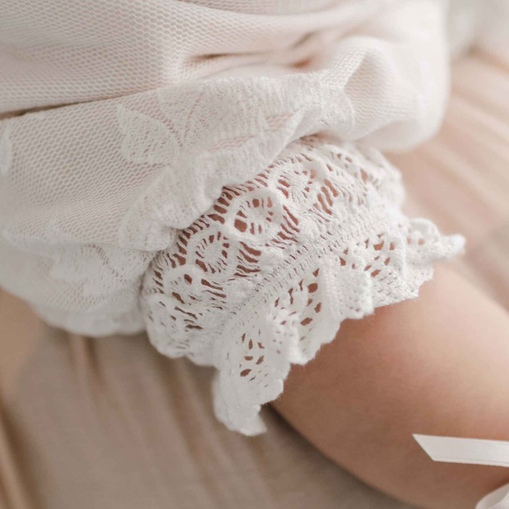 A close-up of the Charlotte Romper features a lace sleeve adorned with intricate, embroidered cotton lace patterns and frills on white fabric. The delicate, swirling designs with a scalloped edge evoke elegance reminiscent of a christening romper.
