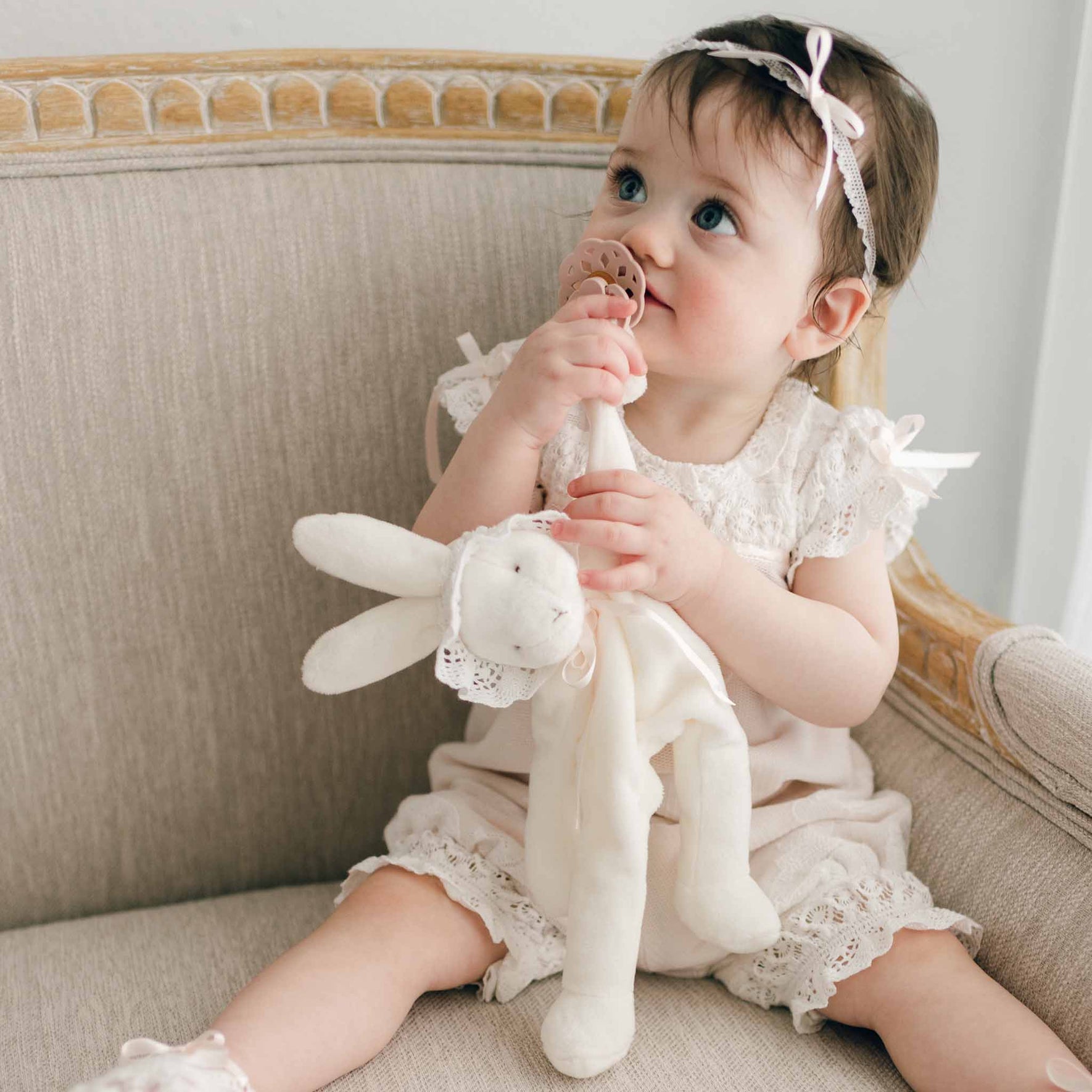 A toddler sits on a beige upholstered chair, adorned in a lace-trimmed outfit with a headband. In one hand, she clutches the Charlotte Silly Bunny Buddy | Pacifier Holder, while gazing slightly upward—perfect for capturing a sweet christening gift memory.