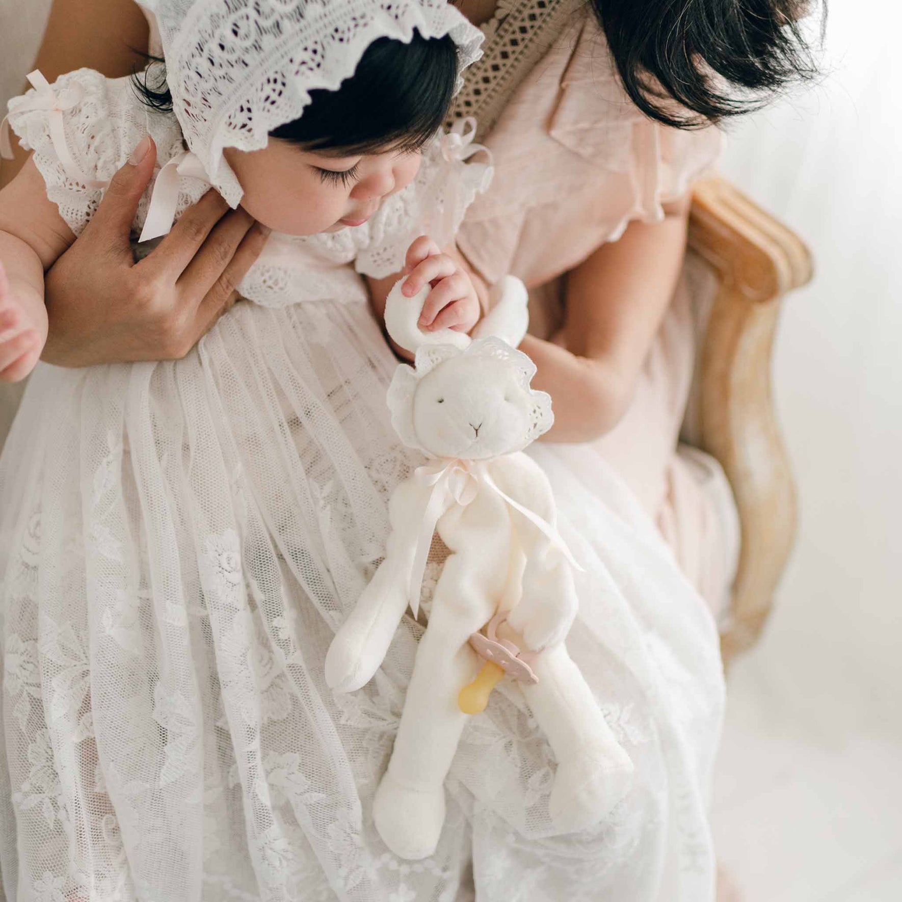 A baby dressed in a white lace dress and bonnet, holding the Charlotte Silly Bunny Buddy | Pacifier Holder, is gently cradled by an adult. The softly lit scene captures the innocence of the moment, with most of the adult's face out of view. This tender image would make a cherished christening gift for any family.
