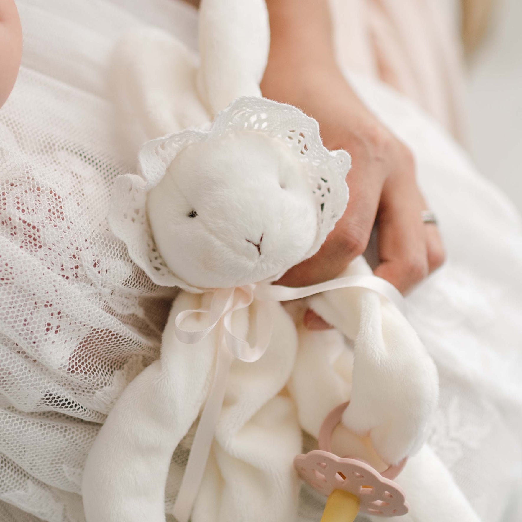 A close-up of the Charlotte Silly Bunny Buddy | Pacifier Holder, a plush white bunny adorned in lace and held by someone. This bunny, ideal for a christening gift, is accessorized with a bonnet and features an attached pacifier clip. The background displays a lace-covered surface.