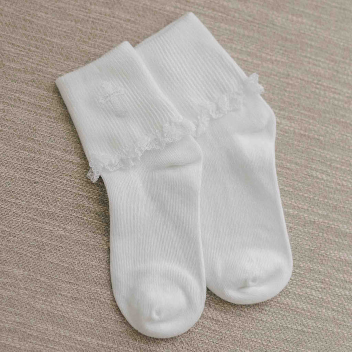 A pair of white Communion socks featuring delicate lace trim and an embroidered cross detail, laid on a neutral fabric surface.