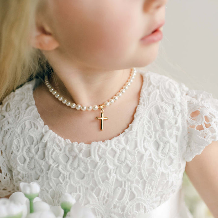 A close-up of a girl's neck adorned with a cream pearl necklace featuring a small gold cross pendant, worn with a white lace dress.