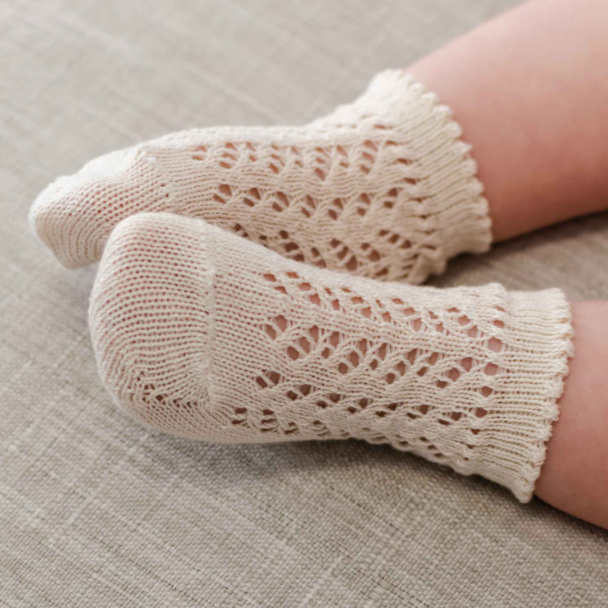Close-up of a baby's feet adorned in Crochet Socks, featuring a cream-colored, intricately crocheted design with a delicate lace pattern. The baby rests on a light-colored fabric surface, enhancing the charm of their cozy ensemble.