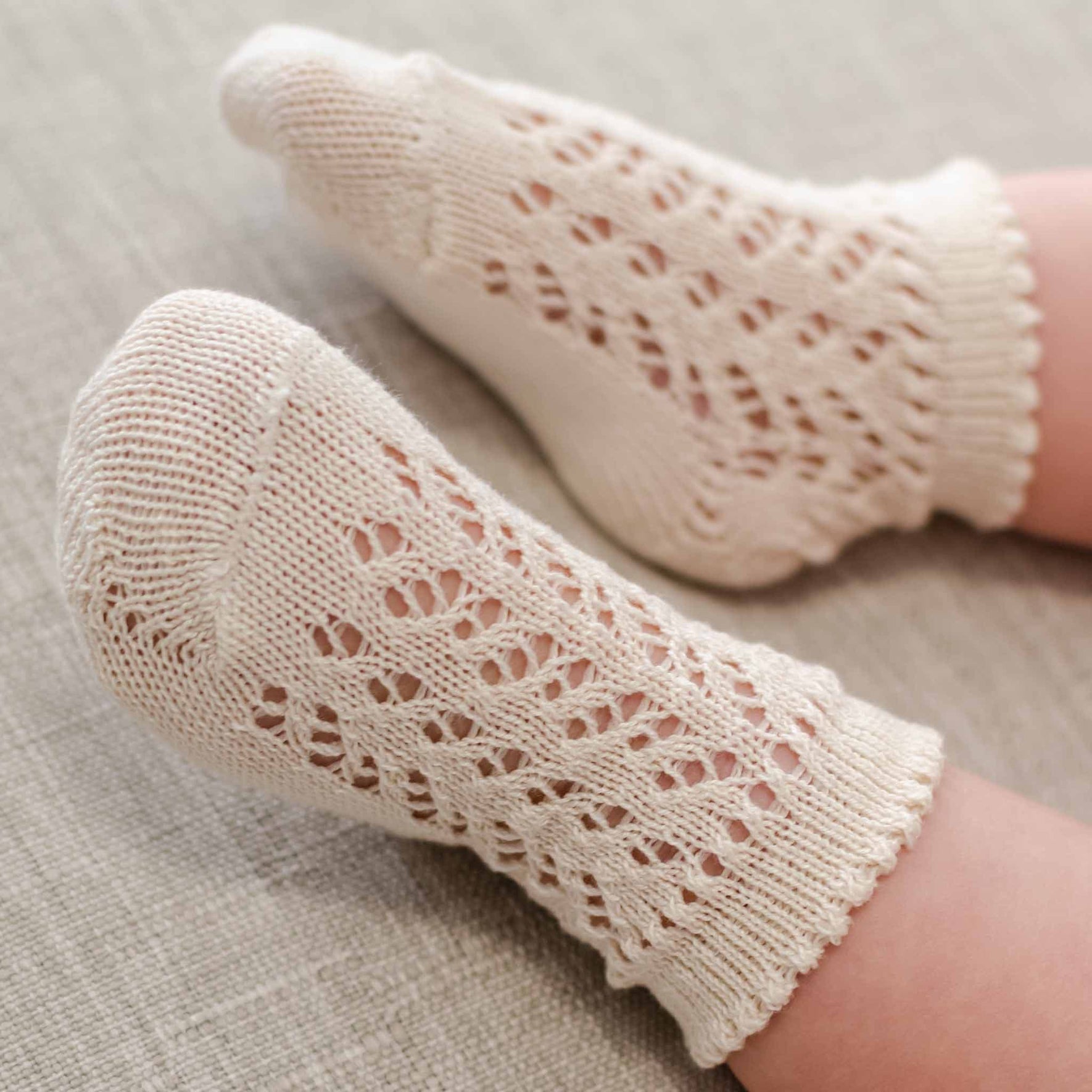 A pair of tiny baby feet is adorned with cream-colored Crochet Socks, showcasing a delicate and intricate openwork design, all set against a light fabric background.
