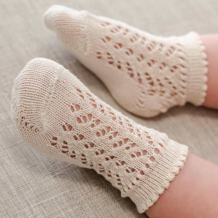 A pair of tiny baby feet is adorned with cream-colored Crochet Socks, showcasing a delicate and intricate openwork design, all set against a light fabric background.