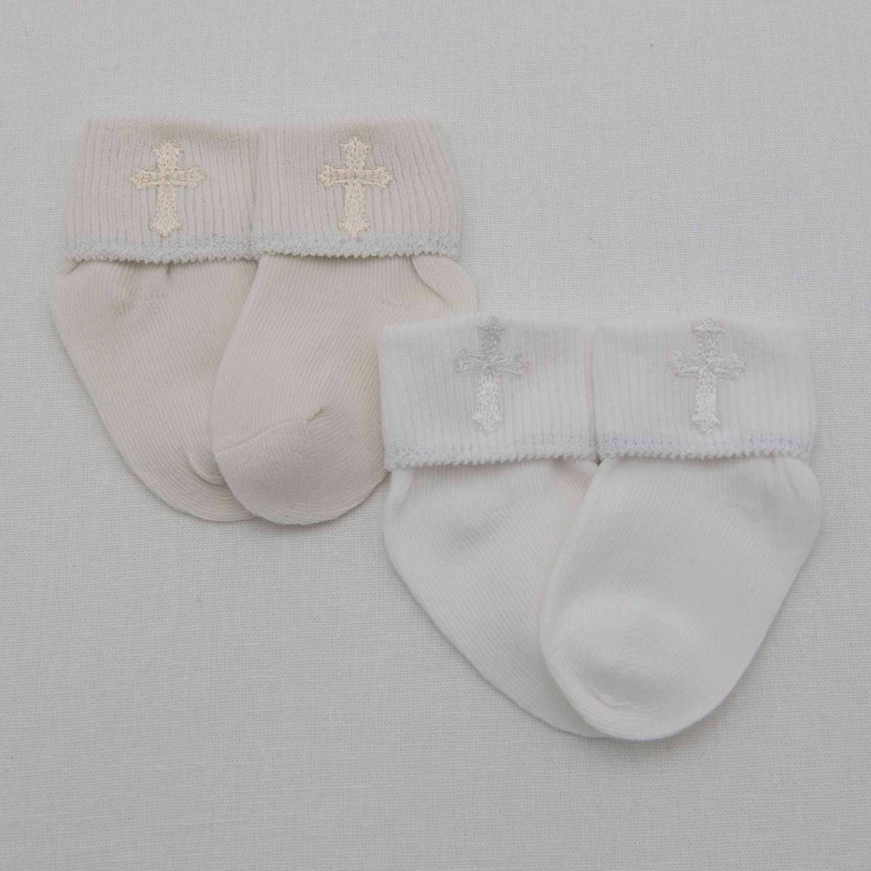 Two sets of Cross Socks are displayed on a plain background. The left pair, made from a comfortable cotton blend, showcases a light beige color with a beige cross design on the folded cuffs. The right set is white with a light gray cross pattern, combining style and comfort for little feet.