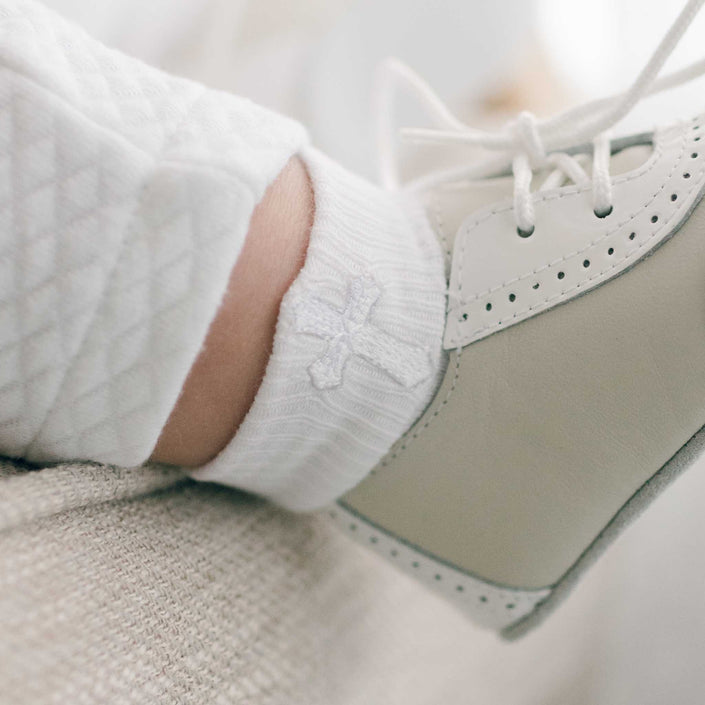 A close-up shot showcases a baby's foot adorned with a white Cross Socks featuring a small cross design, along with a lace-up shoe. The shoe provides a snug fit and is accented by a decorative stitched edge. The soothing, neutral backdrop enhances the cozy feel ideal for little ones.
