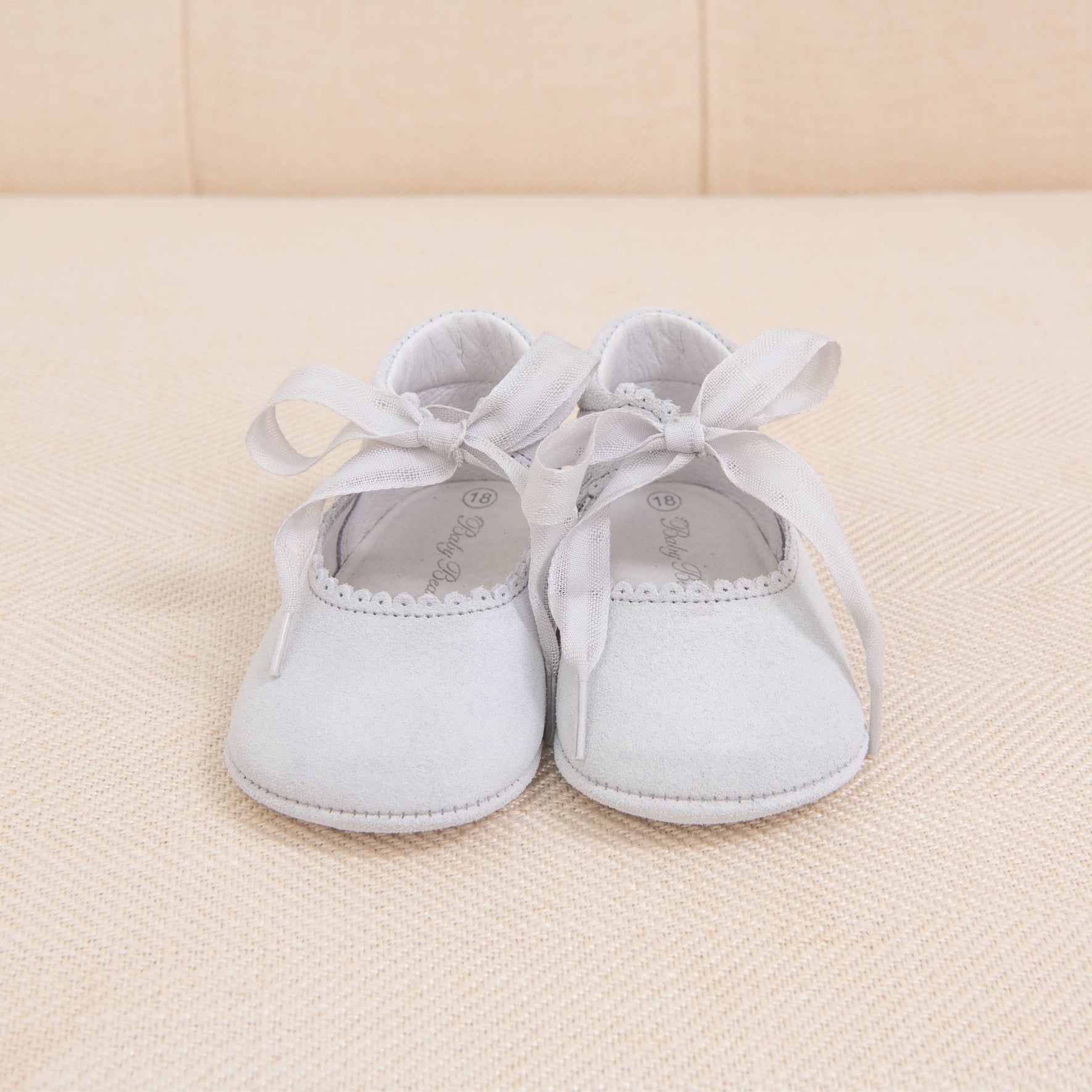 A pair of Dove Grey Suede Tie Mary Janes, handmade in Spain from suede leather with ribbon ties, is placed on a beige fabric surface. The shoes have a decorative edge near the opening and are neatly arranged with their ribbons tied in bows.