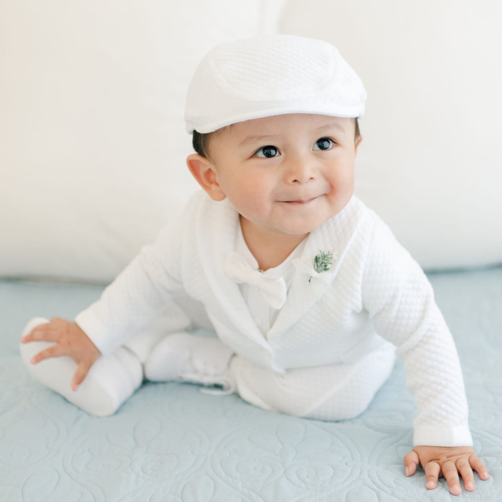 A baby dressed in the Elijah 3-Piece Set, featuring a white christening suit paired with a matching hat, is sitting on a light blue quilt. The baby is looking slightly to the side with a small smile on their face, and white pillows can be seen in the background.