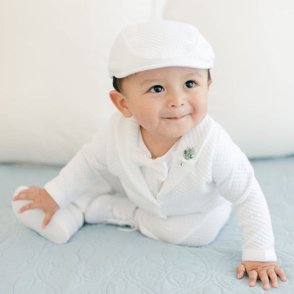 A baby dressed in the Elijah 3-Piece Set, featuring a white christening suit paired with a matching hat, is sitting on a light blue quilt. The baby is looking slightly to the side with a small smile on their face, and white pillows can be seen in the background.