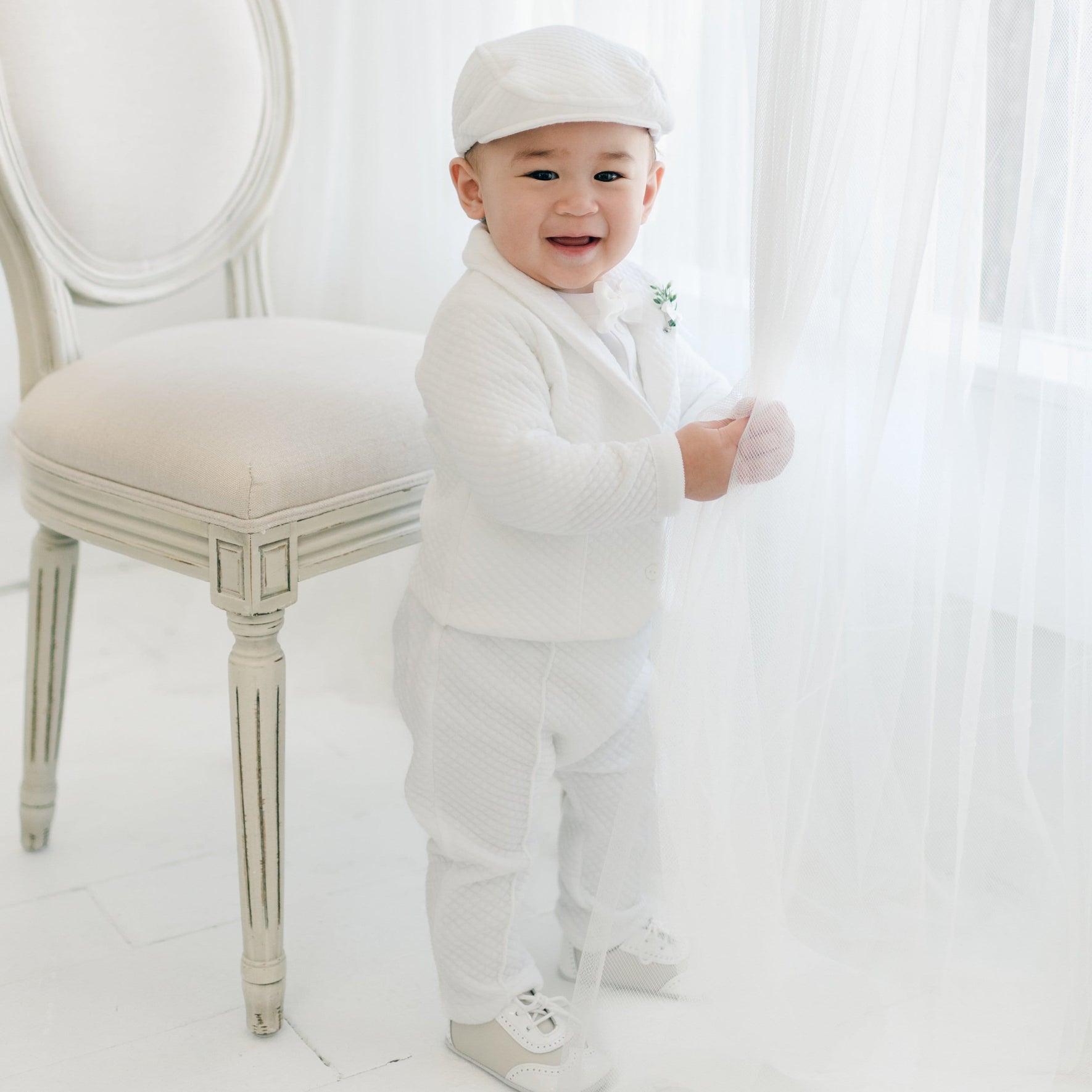 A baby boy stands next to a beige, cushioned chair in a bright room. The baby boy is dressed in the Elijah 3-Piece Set, complete with a jacket, pants, and onesie. Holding onto a sheer white curtain, he smiles at the camera with pure joy. He is also wearing the matching Elijah Newsboy Cap and White Velvet Bowtie & Boutonniere.