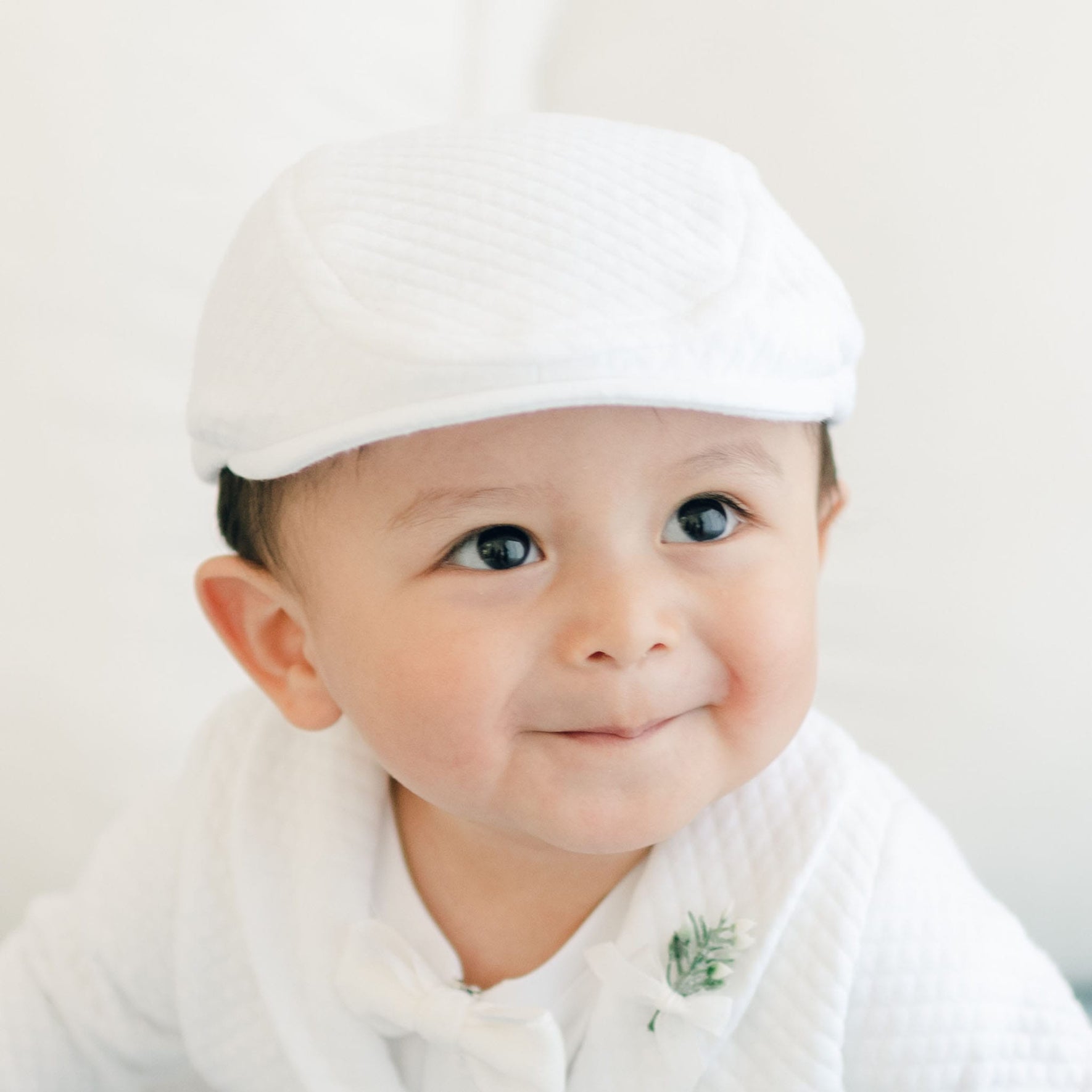 A baby, wearing an Elijah Newsboy Cap and a matching outfit made of white textured cotton, is smiling with a closed-mouth expression. The ensemble includes a bow tie and a small floral accessory pinned to the jacket. The background is white and out of focus.