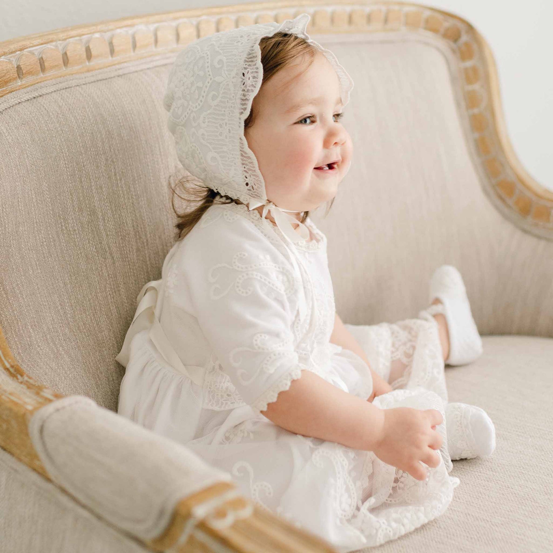 A young child wearing a white lace Eliza Blessing Dress & Headband sits on a beige upholstered chair. The child faces slightly to the right, with a cheerful expression. The chair has light wood accents, and the child is dressed in matching socks made of soft Pima Cotton.