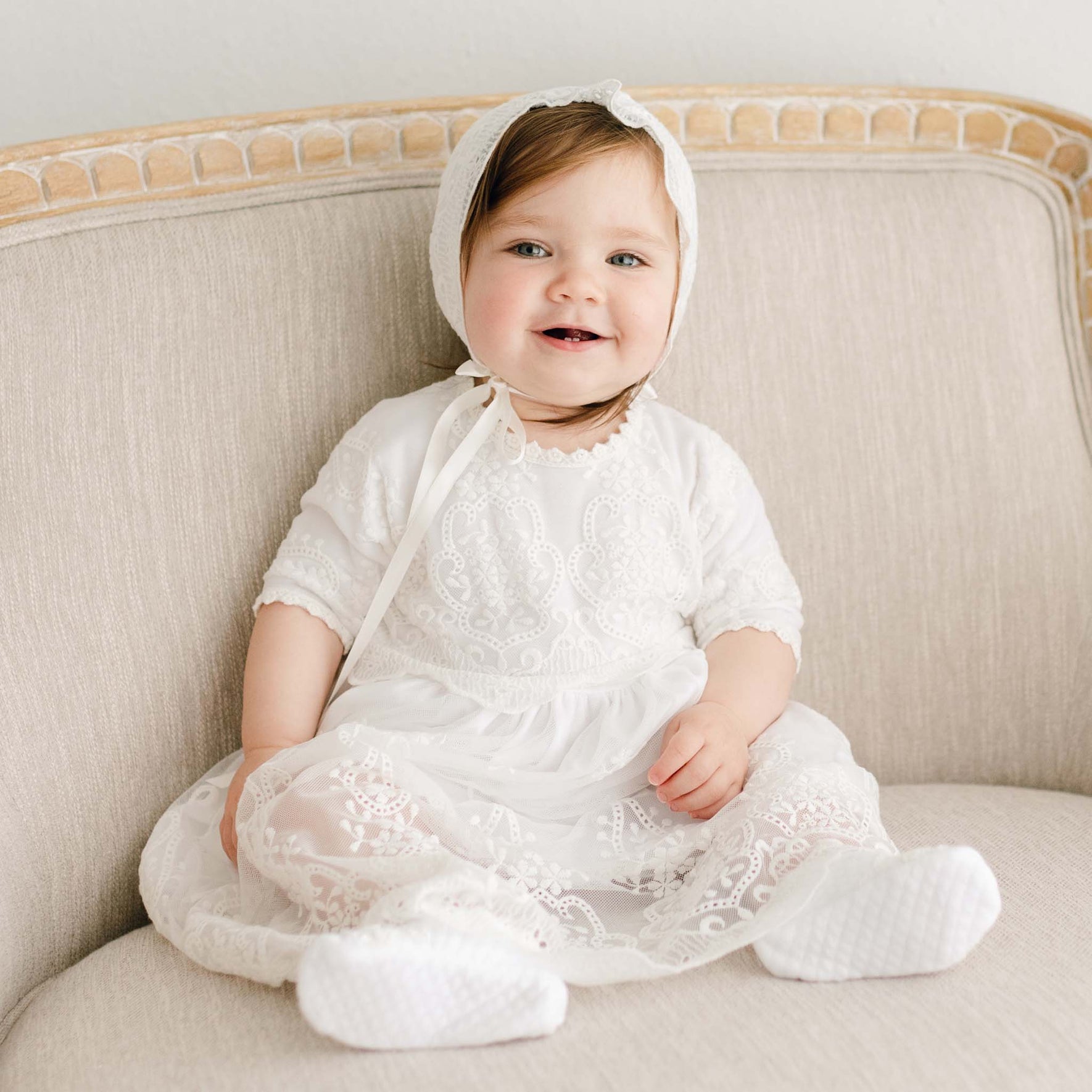 A baby wearing an Eliza Blessing Dress & Headband made of pima cotton is sitting on a beige upholstered chair. The baby is smiling and has one arm resting on the side of the chair, which has a light wood frame with intricate details.