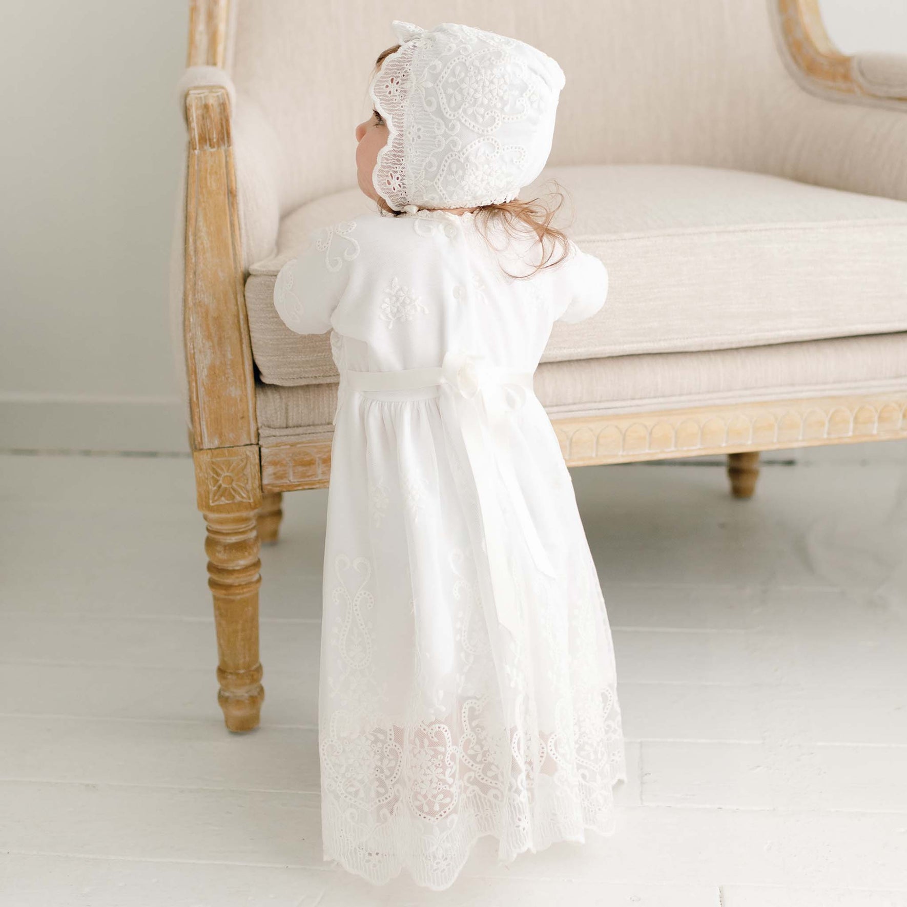 A baby wearing a handmade Eliza Blessing Dress & Headband stands holding onto a beige sofa. The detailed lace patterns on the dress and headband crafted from Pima Cotton are beautifully showcased as the baby faces away. The setting is a bright room with white walls and flooring.
