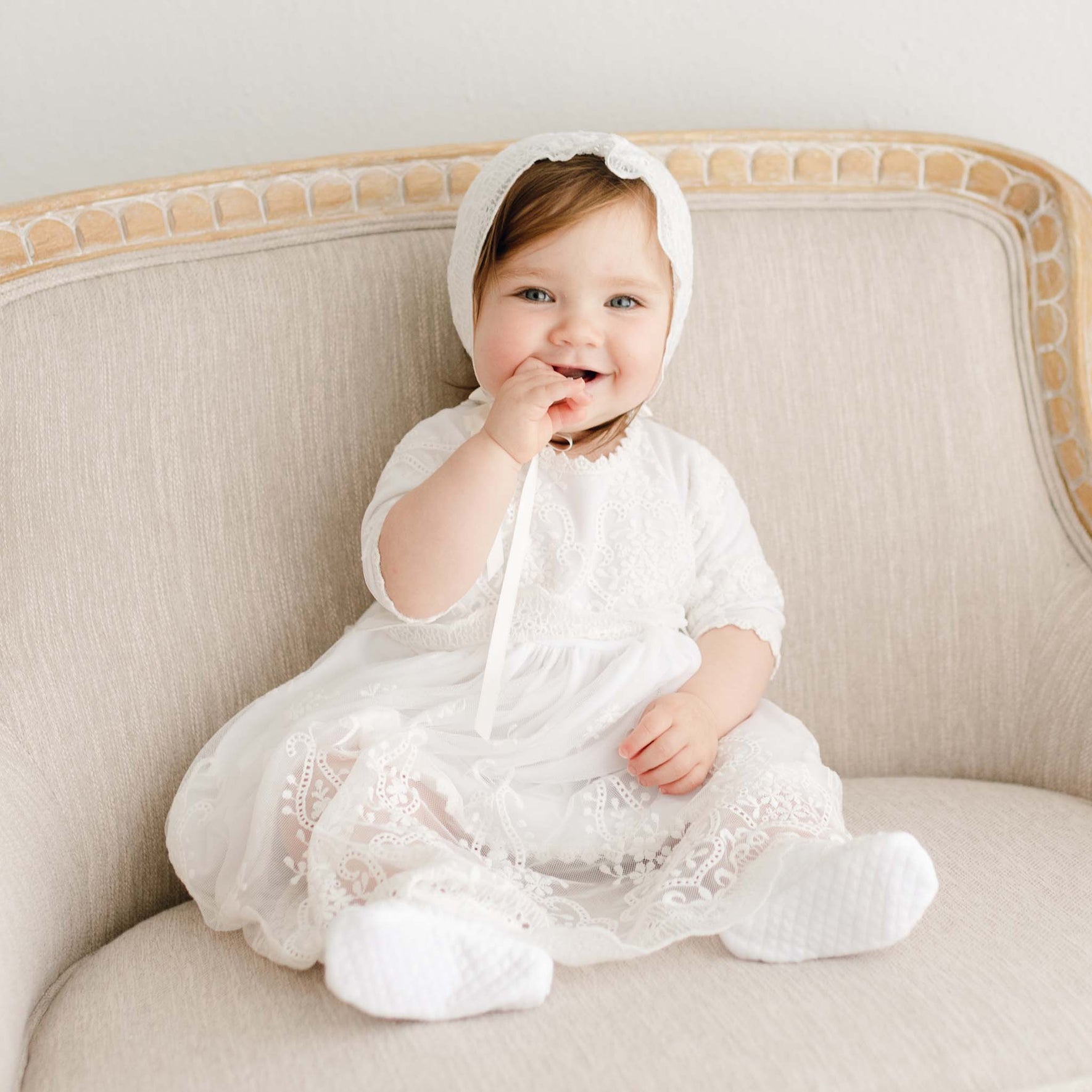 A young child sits on a beige cushioned chair, dressed in a delicate Eliza Blessing Dress & Headband made of Pima Cotton with matching bonnet and white socks. The child is smiling and holding a ribbon up to their mouth, embodying the charm of Handmade USA craftsmanship.