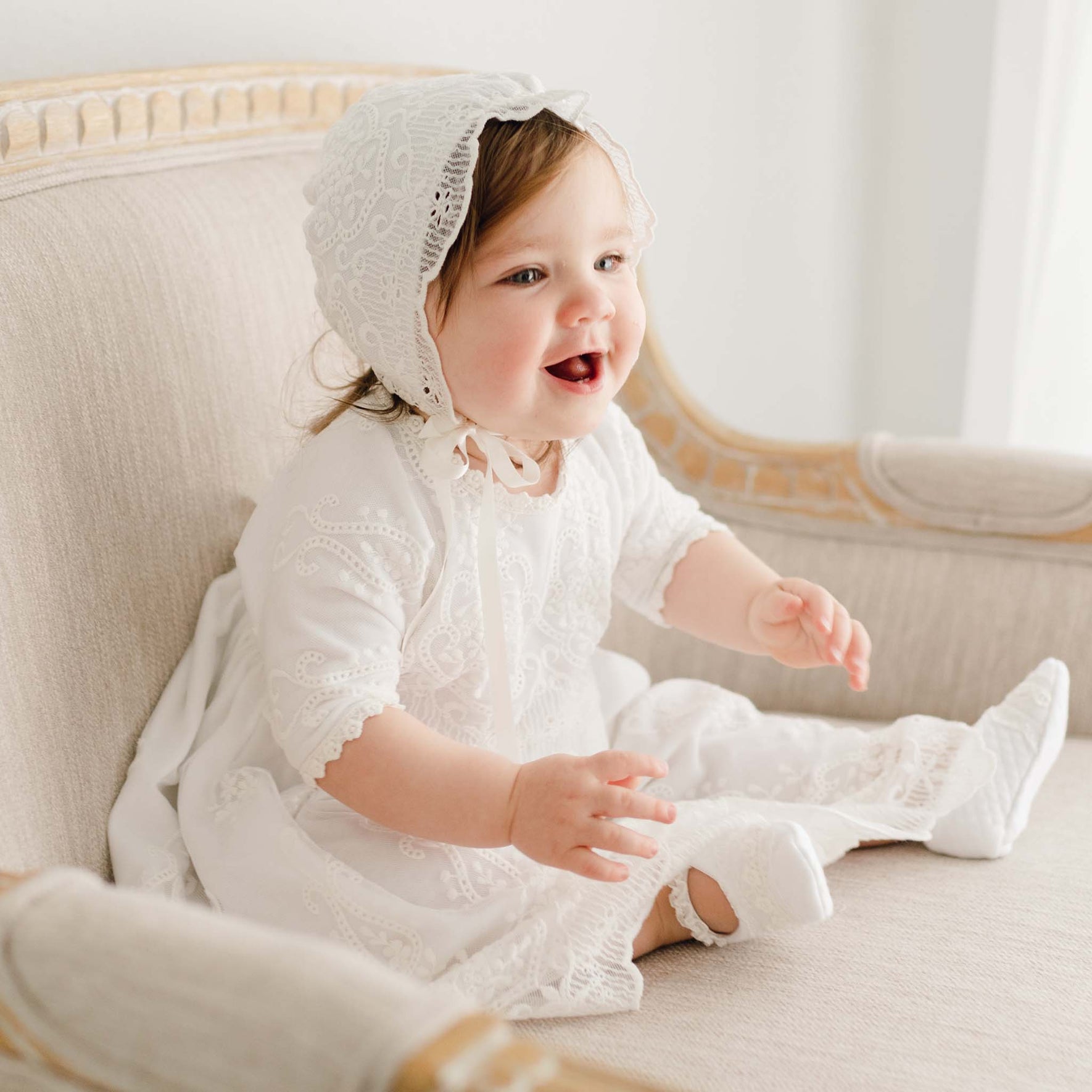 A toddler sits on a beige cushioned chair wearing a delicate Eliza Blessing Dress & Headband made of white lace. The child smiles and extends her arms forward. The background is softly lit and minimalistic, highlighting the beauty of the handmade creation from the USA.