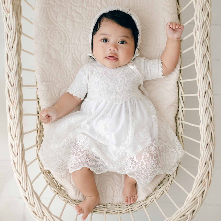 A baby lies in a wicker bassinet, wearing a handmade Eliza Blessing Dress & Headband crafted from soft Pima Cotton. The baby has one arm raised, looking directly at the camera against a light-colored, quilted background.