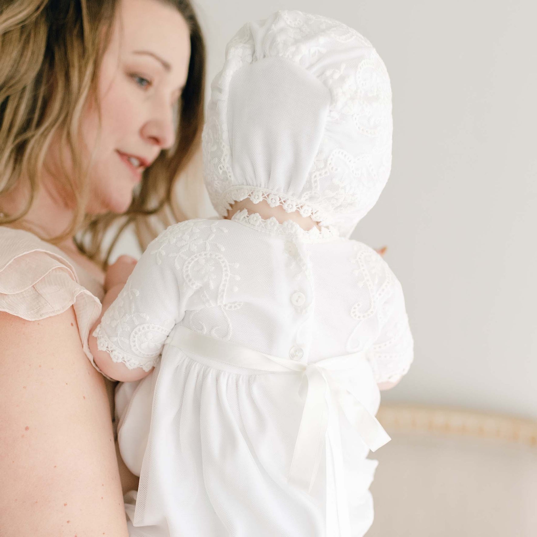 A woman holds a baby dressed in the Eliza Blessing Gown & Bonnet, made from 100% pima cotton, with delicate Christening accessories. The baby is facing away from the camera, and the woman is partially visible, looking at the baby with a soft expression. The background is blurred, focusing on the subjects.