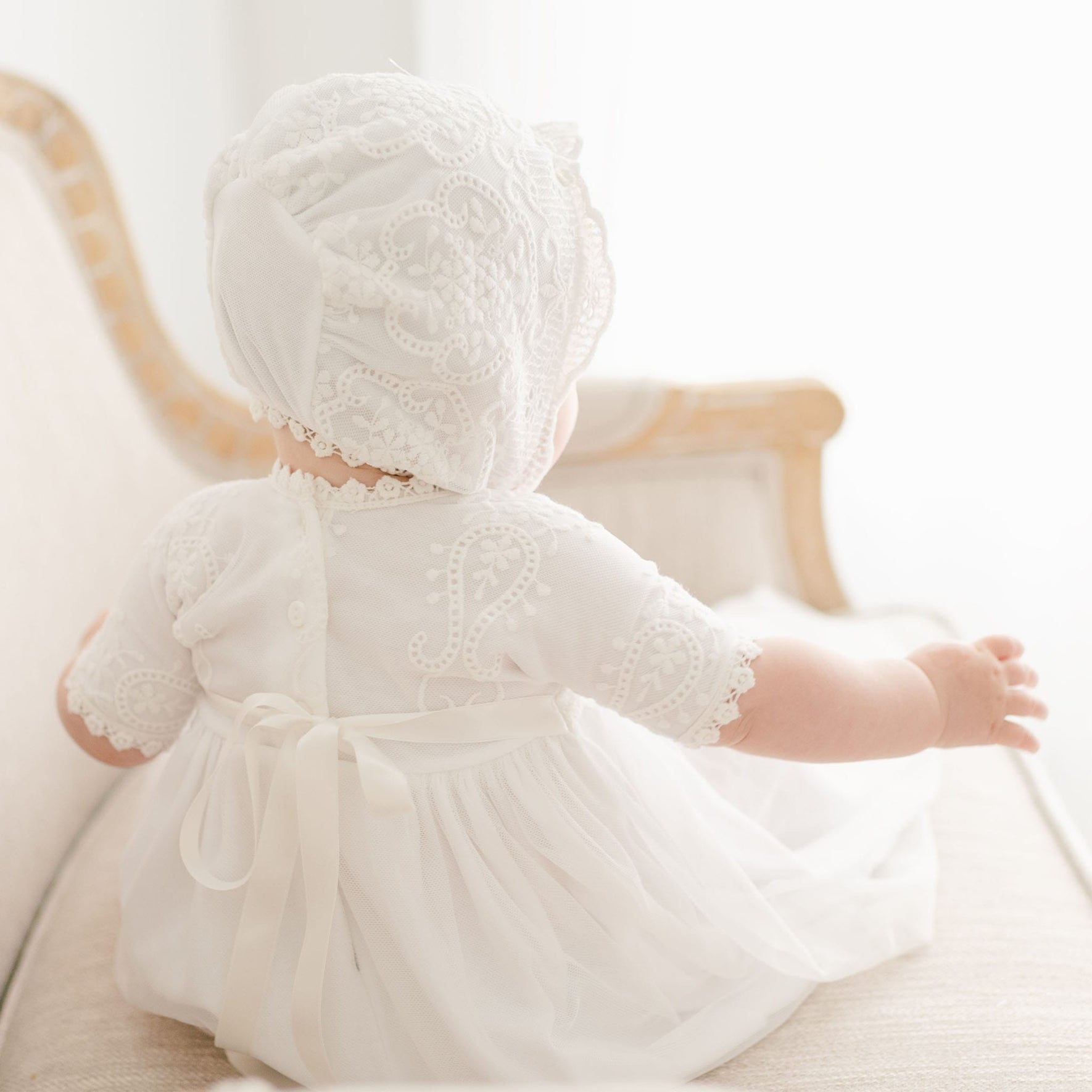 A baby dressed in an Eliza Blessing Gown & Bonnet featuring a white lace bonnet and matching white lace dress sits on a light-colored upholstered chair. The baby is facing away, showcasing the detailed lace and ribbon on the back of the outfit. The background is softly lit, creating a serene atmosphere.