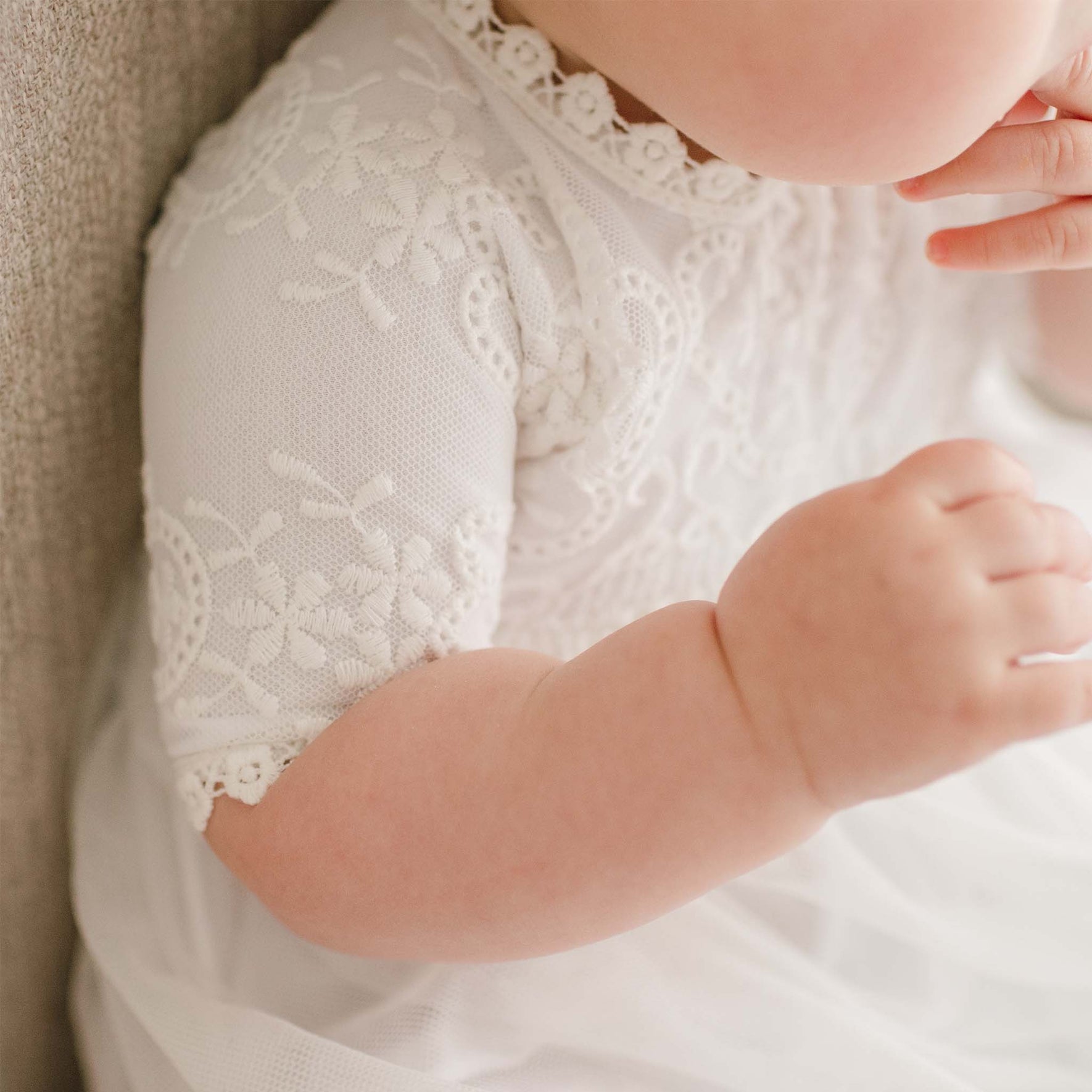 Close-up of a baby wearing an Eliza Blessing Gown & Bonnet crafted from 100% pima cotton. The baby is seated, with one arm bent and hand near the face, and the other arm resting on their leg. The intricate lace details of the dress are clearly visible. The background is neutral and out of focus.