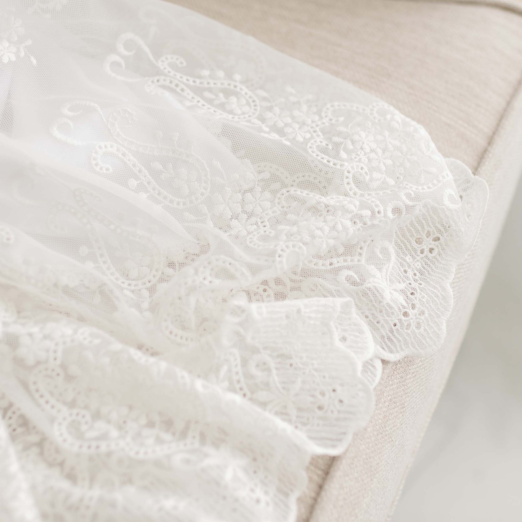 Close-up of a sheer white lace fabric with intricate floral and paisley patterns laid over a cream-colored upholstered surface. The lace, often used in Christening accessories like the Eliza Blessing Gown & Bonnet, features detailed embroidery showcasing delicate designs and fine craftsmanship.