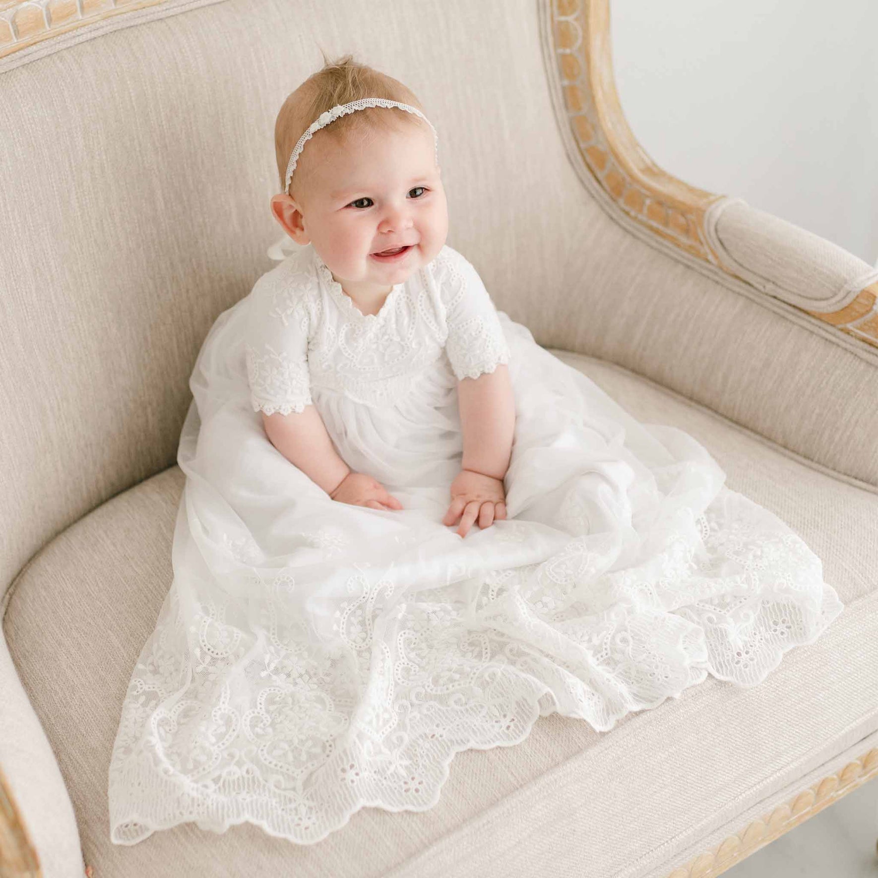 A baby wearing the Eliza Blessing Gown & Bonnet made of 100% pima cotton and a delicate headband sits on a beige upholstered chair with wooden trim, smiling and looking slightly to the side. The dress features intricate lace detailing, perfect for showcasing Christening accessories.