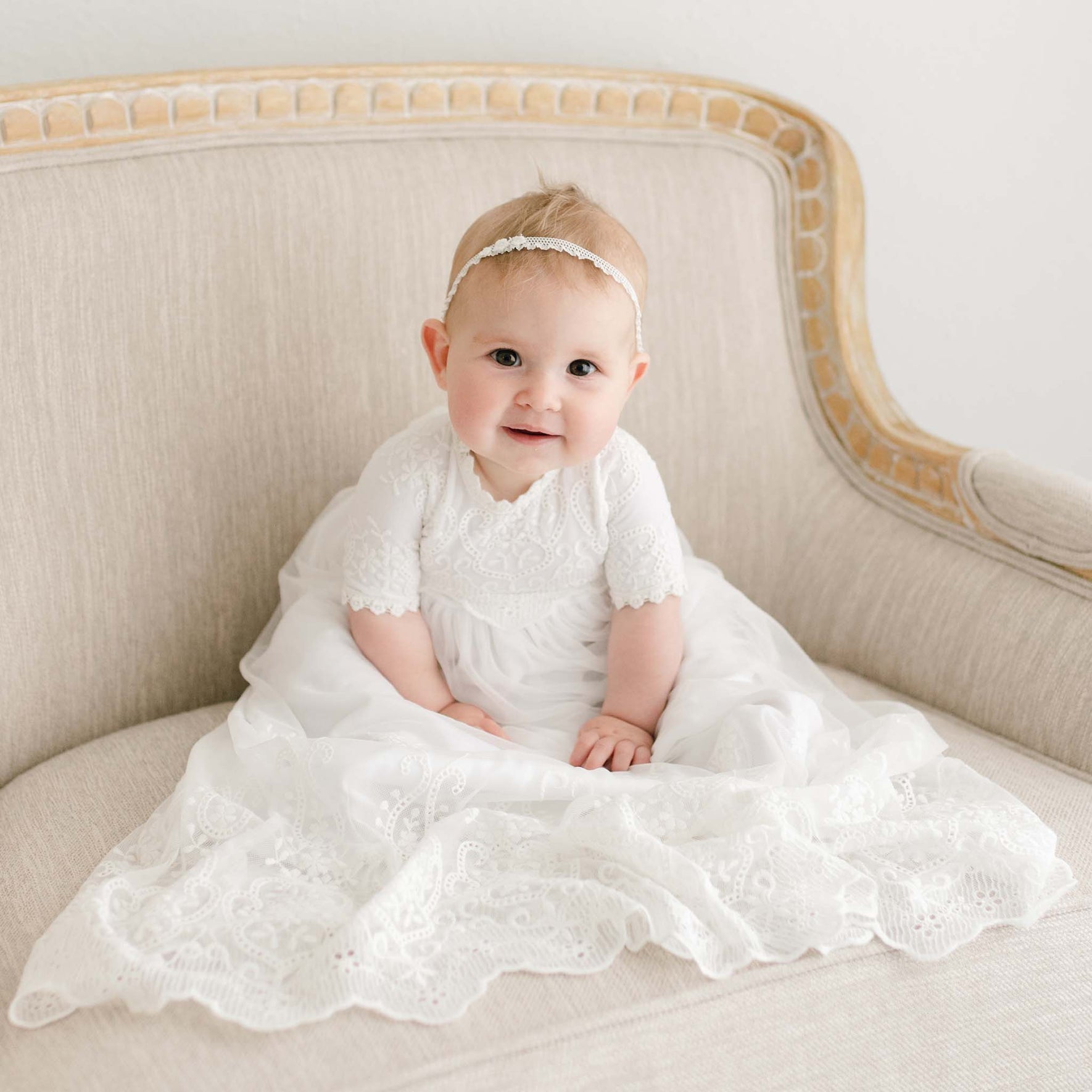A baby dressed in an Eliza Blessing Gown & Bonnet and headband sits on a beige upholstered chair. The baby is looking at the camera with a slight smile. The background is plain white, creating a clean and minimalistic setting.