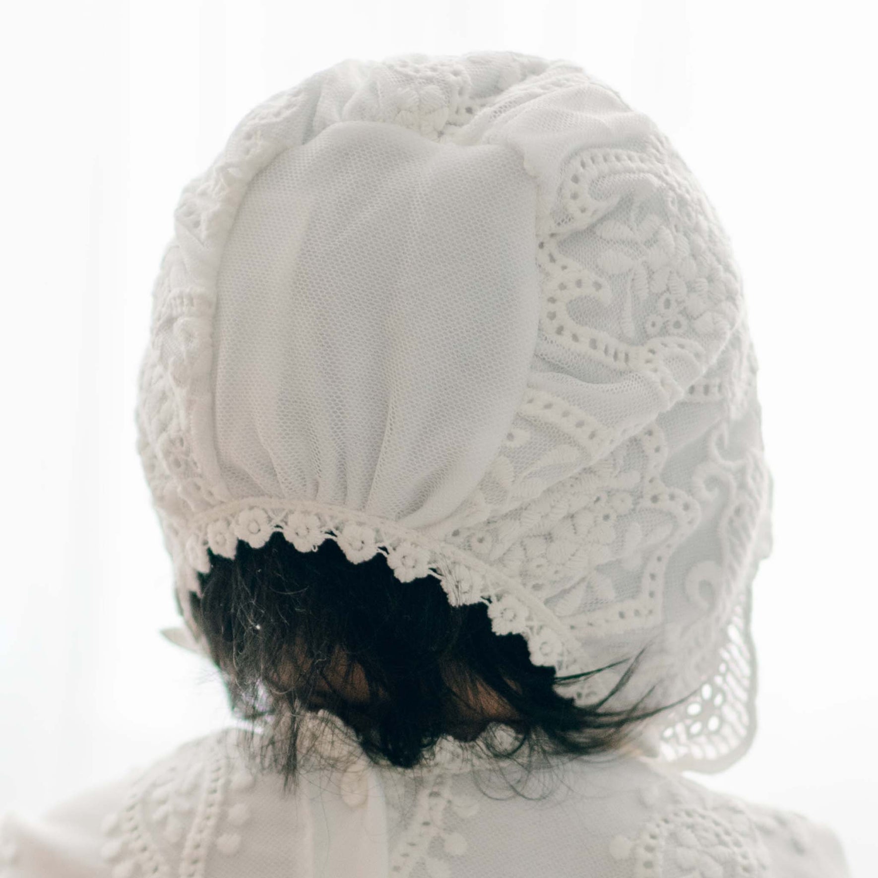 A close-up of the back of the Eliza Christening Bonnet as featured on a baby girl with dark hair. The close-up showcases the intricate lacework on the bonnet.