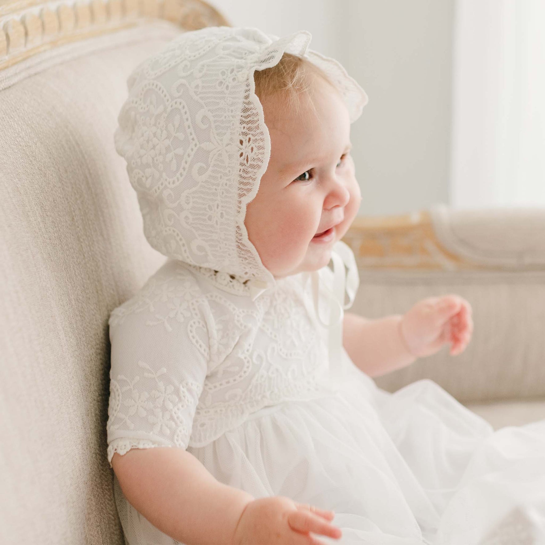 A smiling baby in the Eliza Christening Bonnet and matching baptism gown sits on a upholstered beige chair and looks to the side with a bright expression.