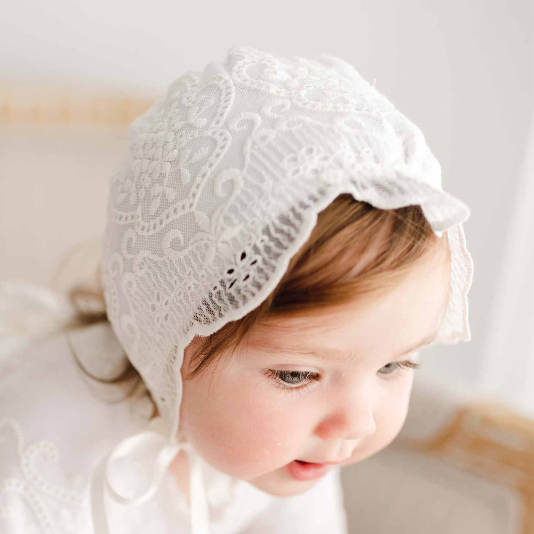 A toddler with light brown hair wears the Eliza Christening Bonnet and matching baptism dress, gazing downward with a gentle expression and slight smile.