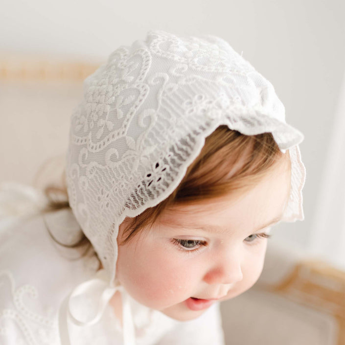 A toddler with light brown hair wears the Eliza Christening Bonnet and matching baptism dress, gazing downward with a gentle expression and slight smile.