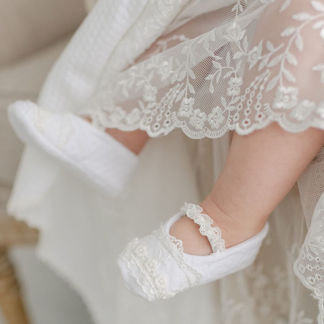 A close-up shot captures a baby's feet in Eliza Booties, embellished with intricate white floral lace. The baby is attired in a cream-colored dress adorned with lace, creating an air of softness and delicacy.