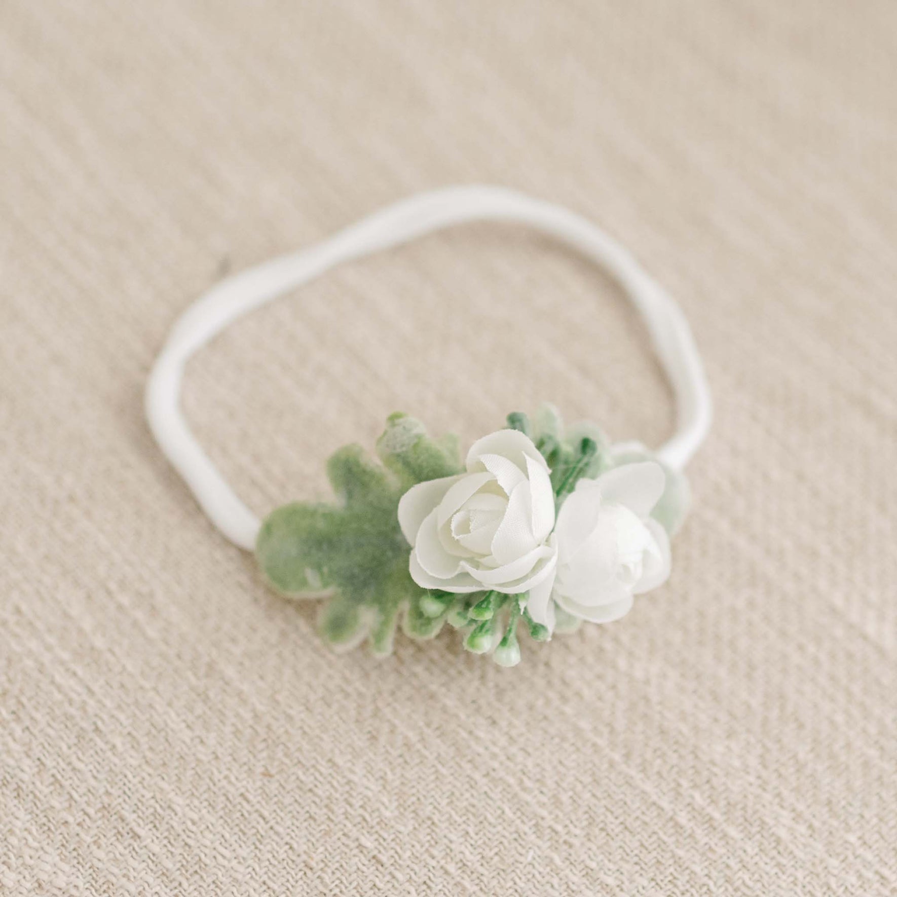 The Eliza Flower Headband, featuring delicate ivory flowers and lush green leaves, rests gracefully on a beige textured fabric background, highlighting its elegant design.