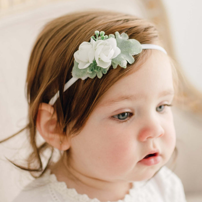 A young child with light brown hair is wearing the Eliza Flower Headband, which is adorned with ivory flowers and greenery on a soft nylon band. The child, shown from the shoulders up in a white outfit, appears to be looking off-camera.