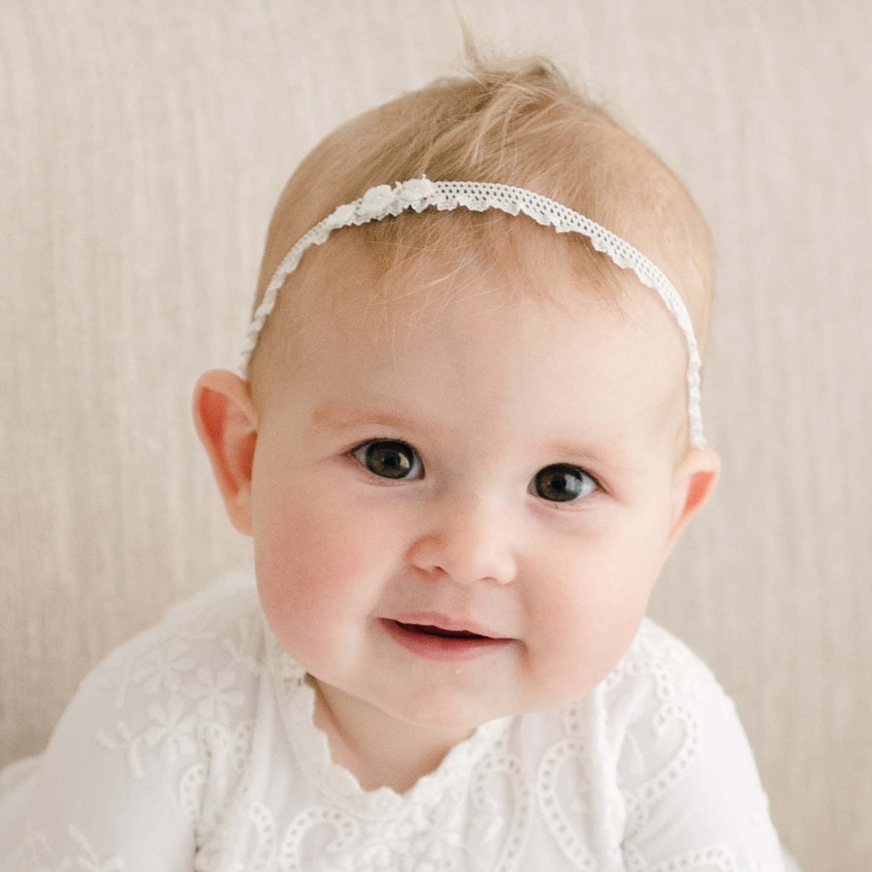 A baby wearing an elegant white lace christening gown with floral appliques and an Eliza Lace Baby Headband smiles as they gaze directly at the camera, set against a neutral beige background.
