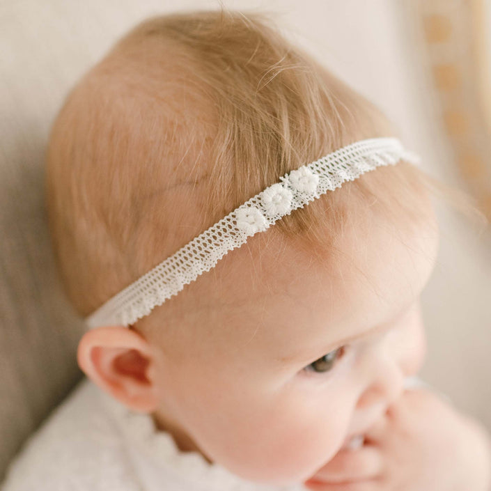 A baby with light hair wears a delicate white Eliza Lace Baby Headband, featuring small floral appliques, against a neutral background. The baby has a thoughtful expression and slightly sucks their thumb.