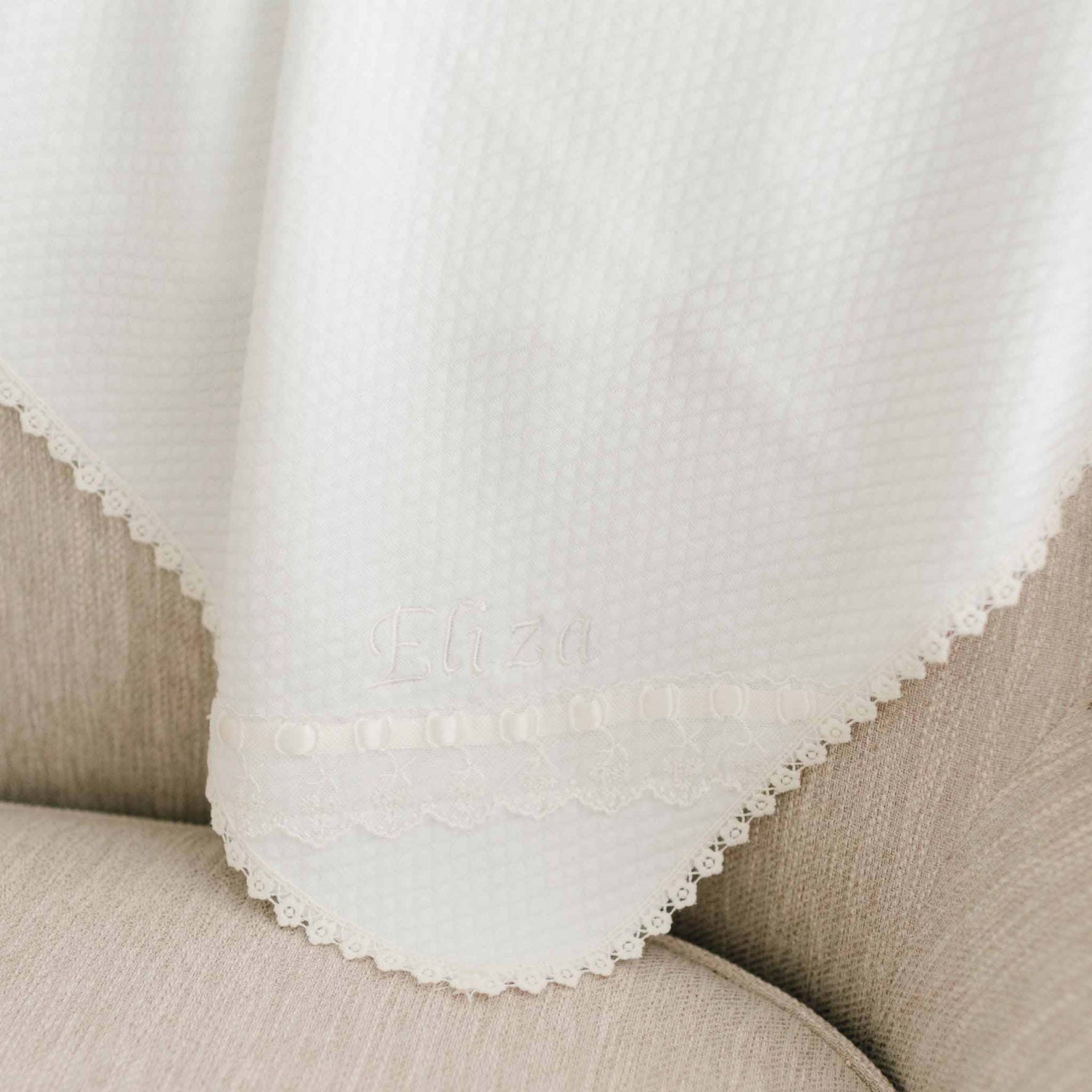 A white textured fabric with floral lace trim is draped over a beige upholstered surface. The Eliza Personalized Blanket, featuring personalized embroidery with the name "Eliza" in light thread near the lower edge, exudes a unique and delicate charm.