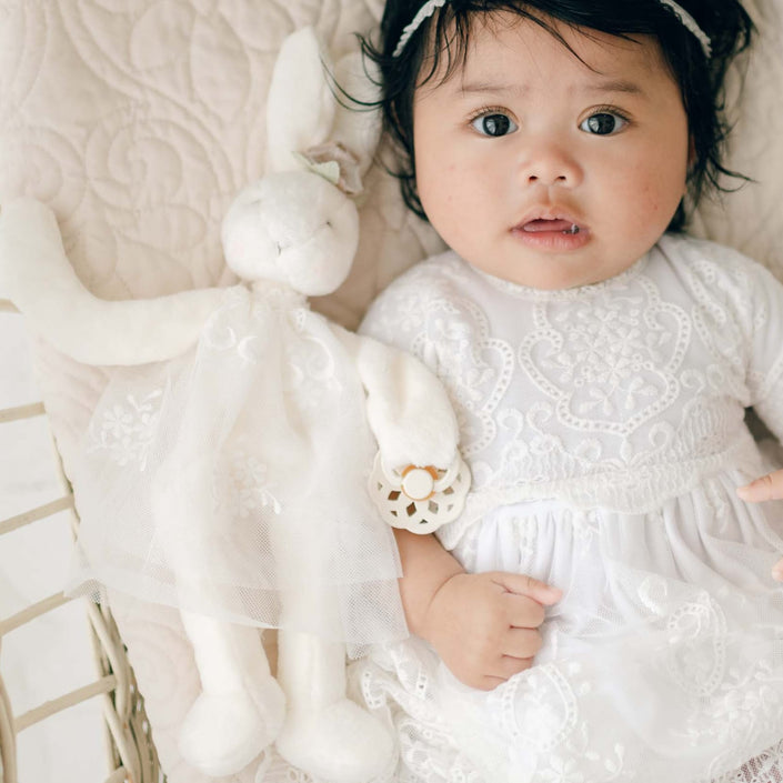 A baby dressed in a white lace outfit lies on a quilted blanket in a crib, holding an Eliza Silly Bunny Buddy and looking up with wide eyes. A thin lace headband adorns the baby's head, and beside them is an intricate Eliza Silly Bunny Buddy | Pacifier Holder.