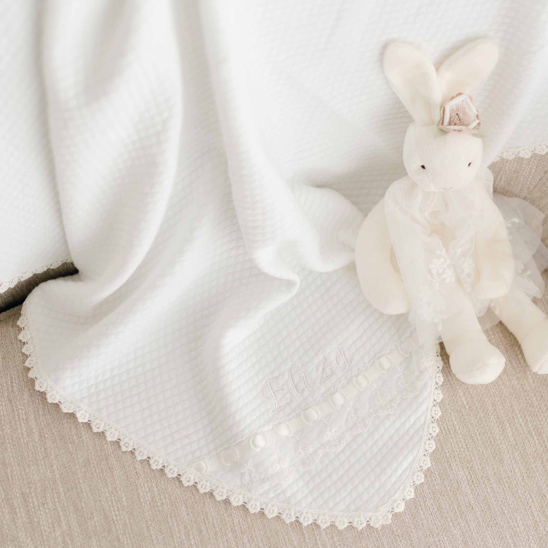 A white blanket with lace trim draped on a beige surface. Next to the blanket sits an Eliza Silly Bunny Buddy | Pacifier Holder wearing a lace headband and a lace ruffle on one ear. The word "Eliza" is embroidered on the blanket.