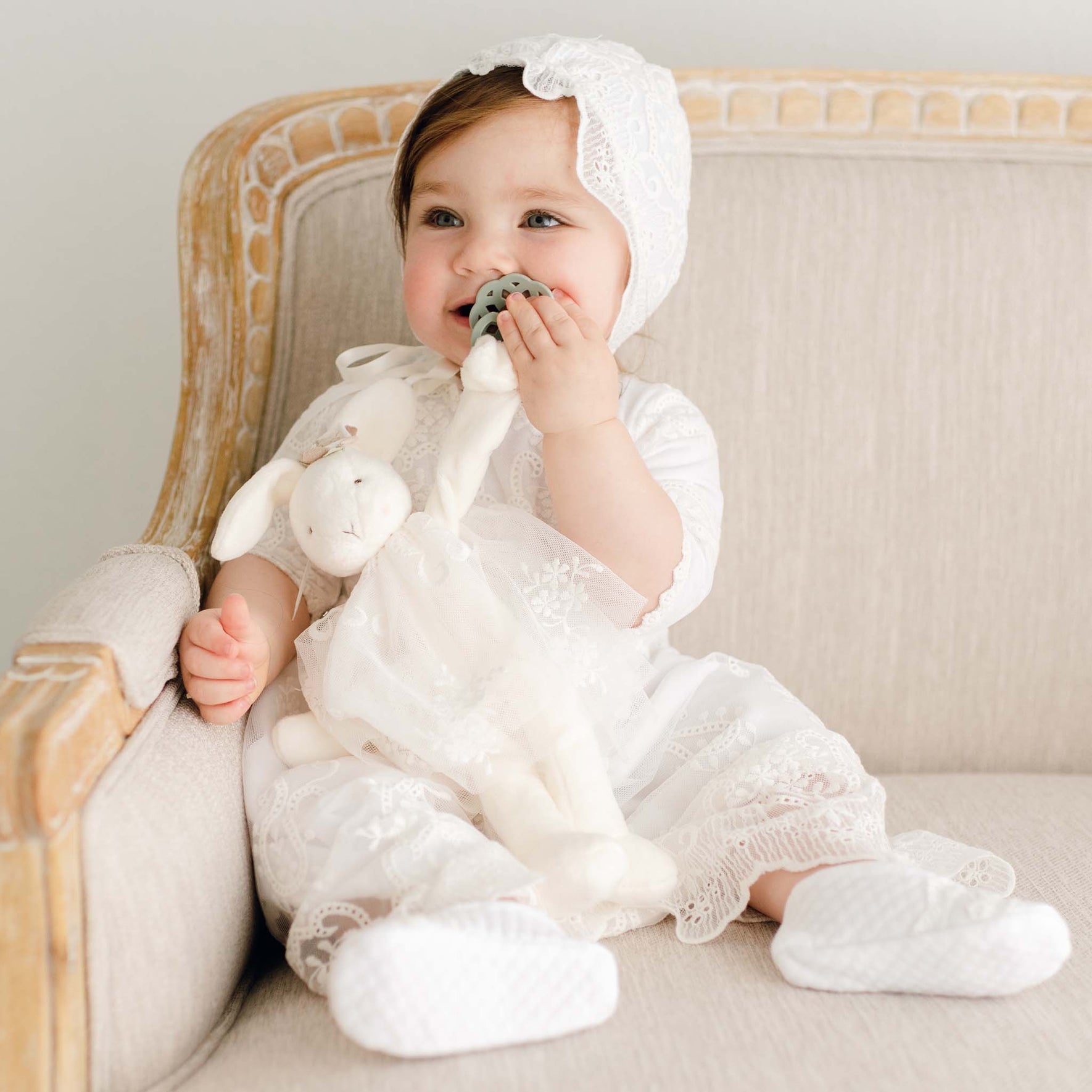 A baby in a white lace outfit and bonnet sits on a beige cushioned chair, holding an Eliza Silly Bunny Buddy | Pacifier Holder and chewing on a teething toy.