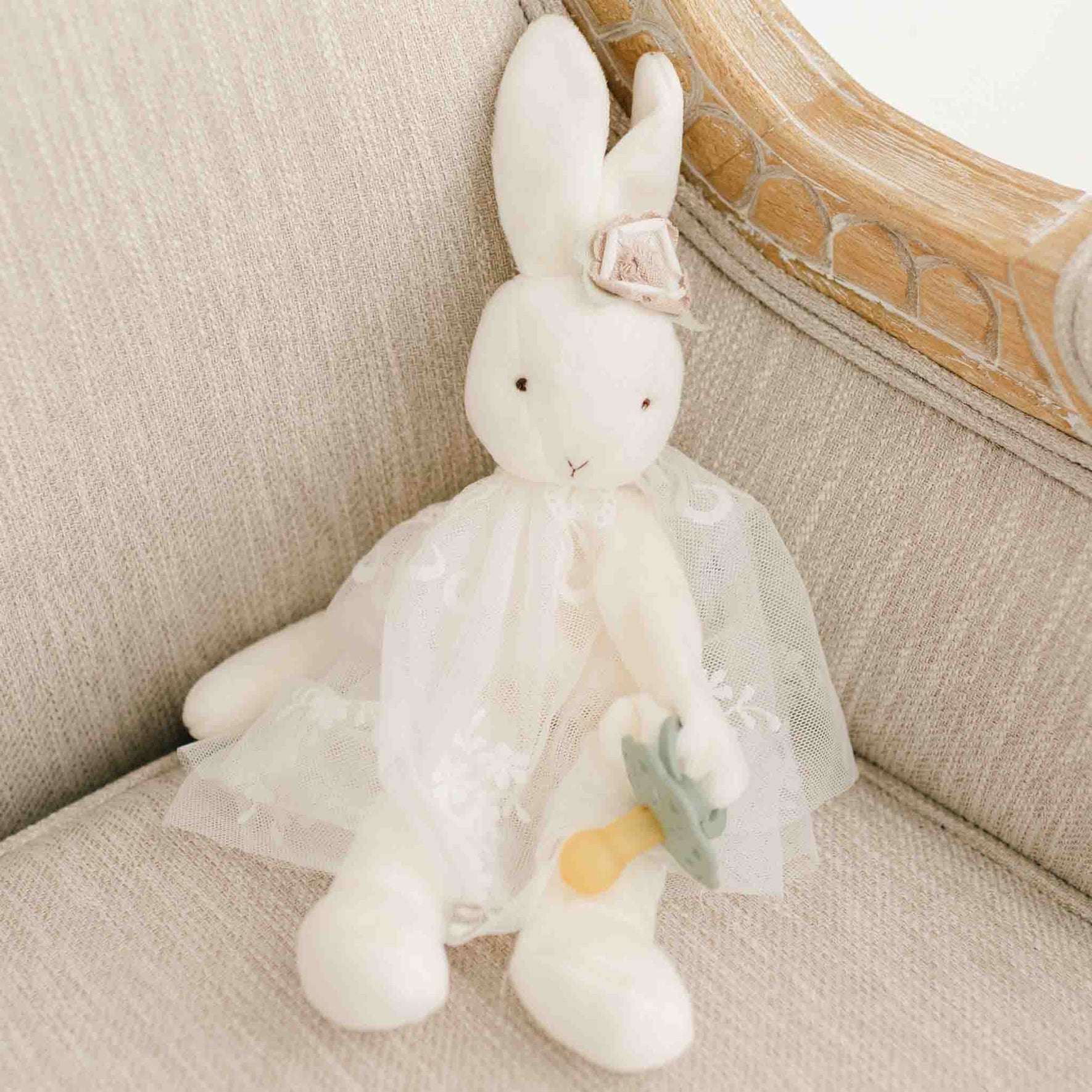 A white plush Eliza Silly Bunny Buddy | Pacifier Holder sits on a beige couch. The bunny is dressed in a white lace dress and has a small bow on one of its ears. It holds a small blue-grey toy umbrella in one hand, making it the perfect baby accessory.