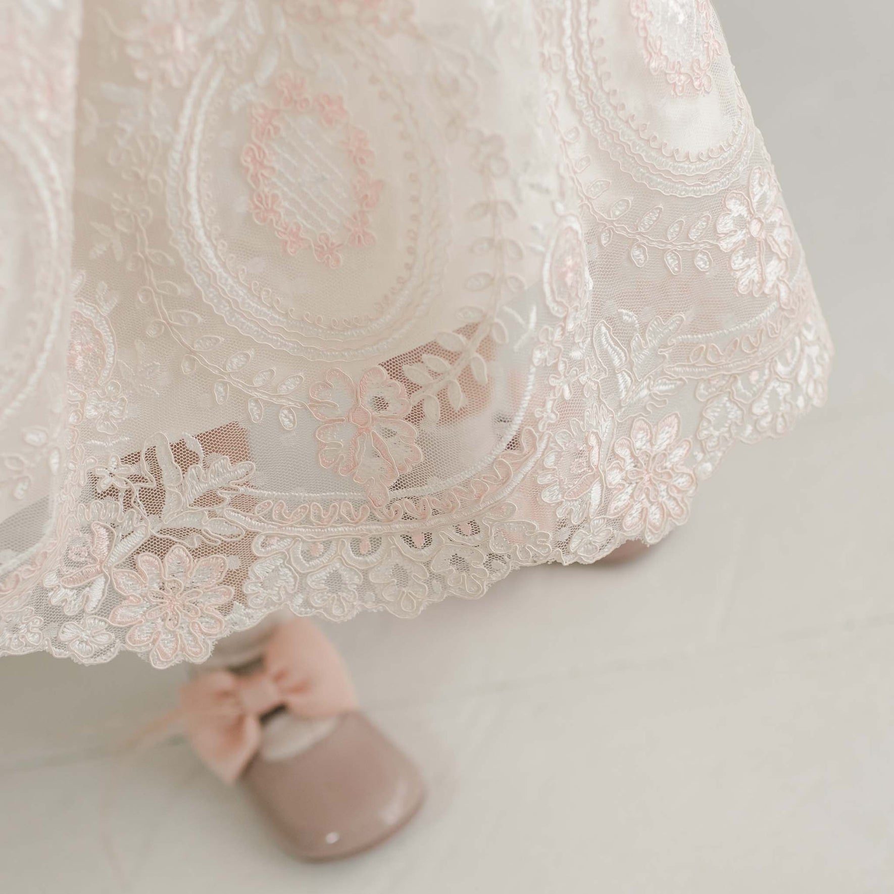 A close-up view of the hem of the Elizabeth Christening Dress reveals cream-colored lace adorned with pink floral embroidery. Below the hemline, a pair of shiny taupe shoes featuring pink bows stand on a white surface. The vintage lace detailing imparts an elegant, handmade touch to the ensemble.