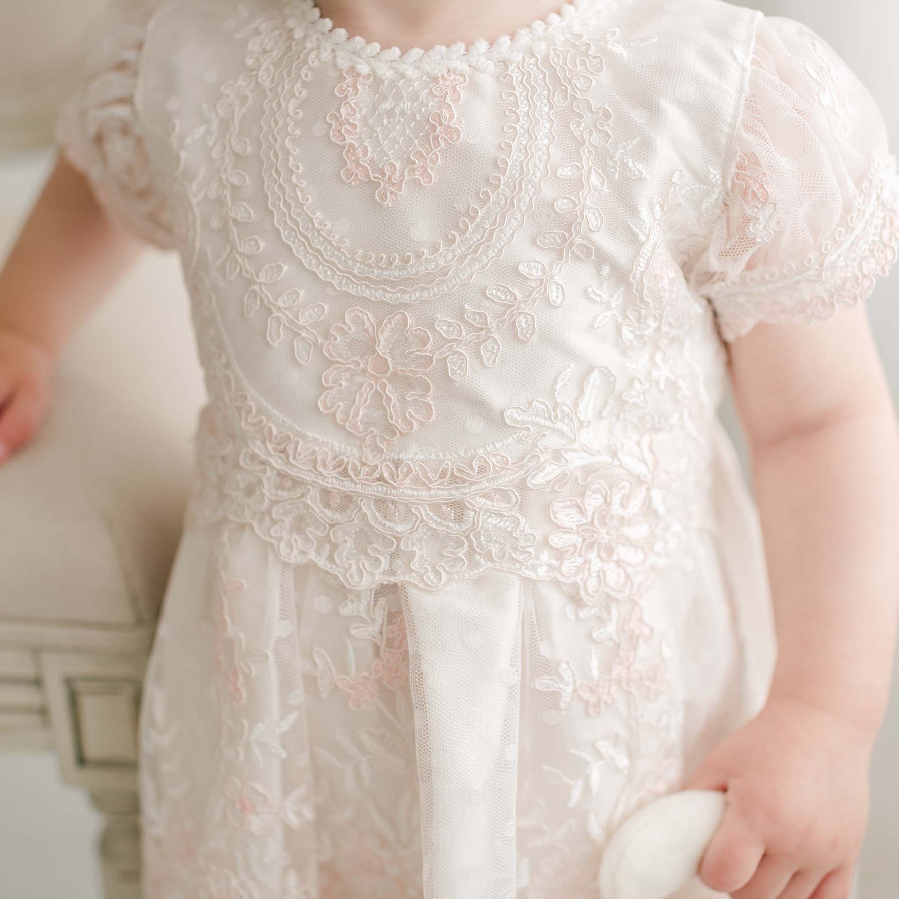 A young child wears the Elizabeth Christening Dress, an intricately embroidered white Baptism gown adorned with floral patterns and lace details. The handmade dress, crafted from luxurious silk Dupioni, is beautifully complemented by a small white object in the child’s right hand. The photo highlights the upper body and showcases the exquisite dress details.